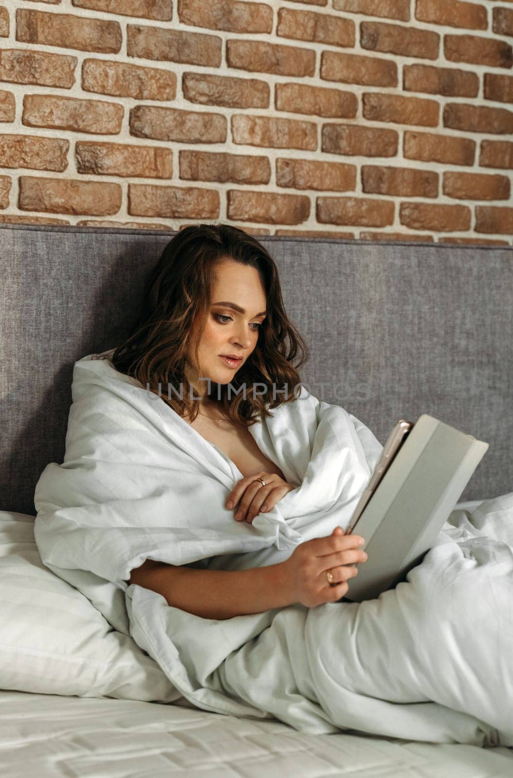 A young woman lies in bed in the morning, watches the news on her tablet. Vertical frame.