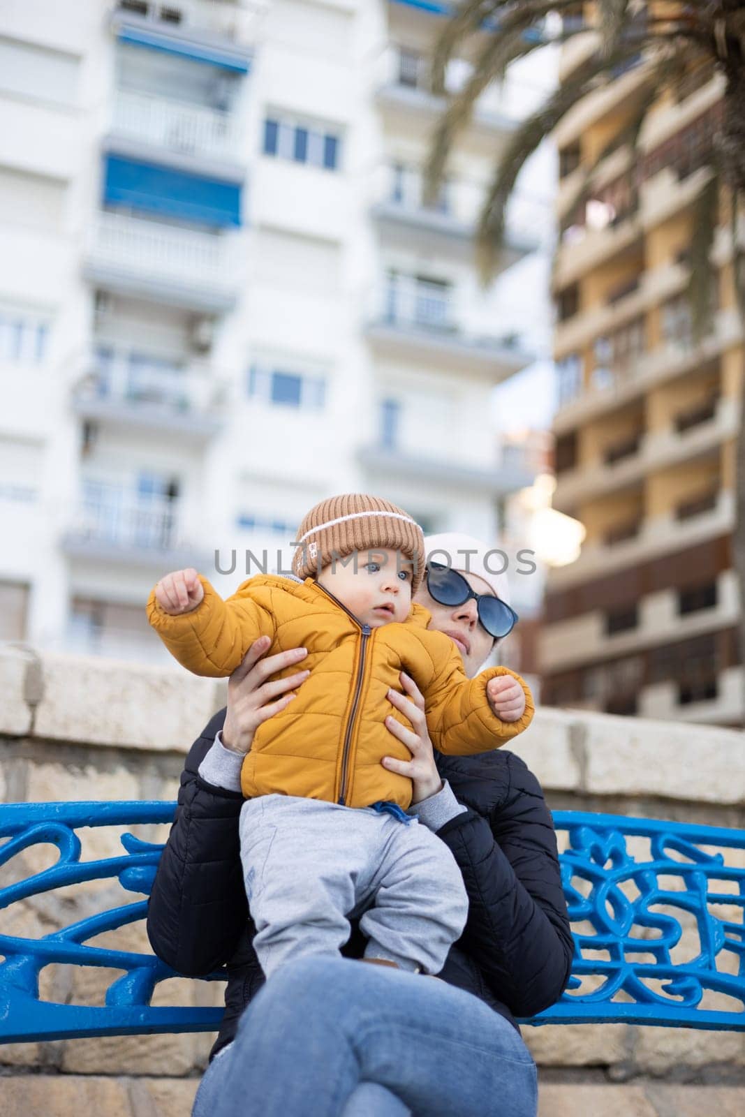 Young mother with her cute infant baby boy child on bench in city park