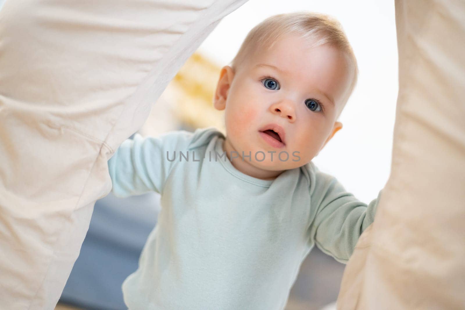 Portrait of adorable curious infant baby boy child taking first steps holding to father's pants at home. Cute baby boy learning to walk