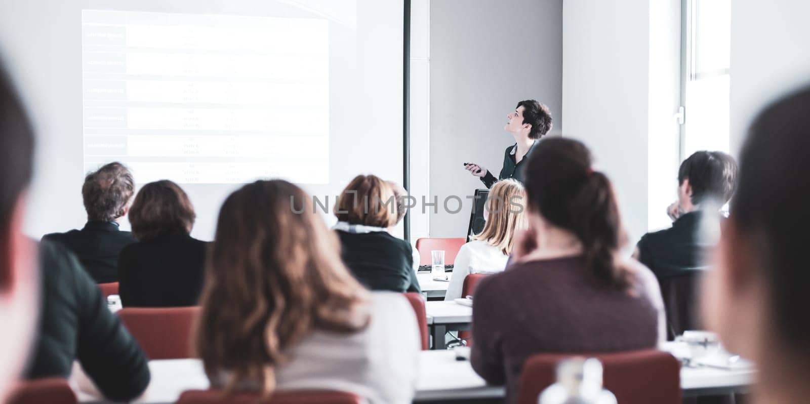 Female speaker giving presentation in lecture hall at university workshop . Participants listening to lecture and making notes. Scientific conference event.