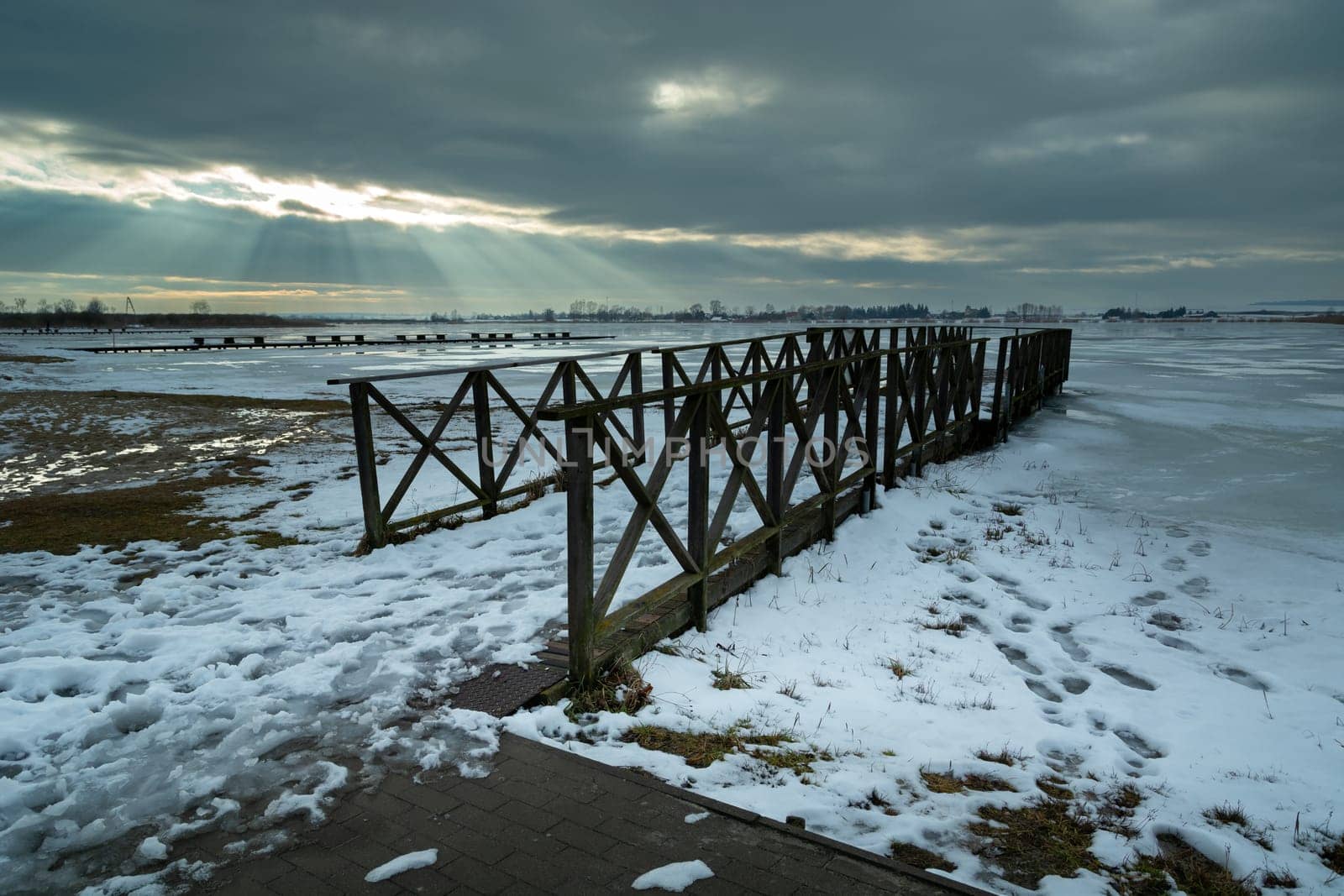 Winter view of a wooden pier and a frozen lake, sunbeams in the clouds, Zoltance, Poland
