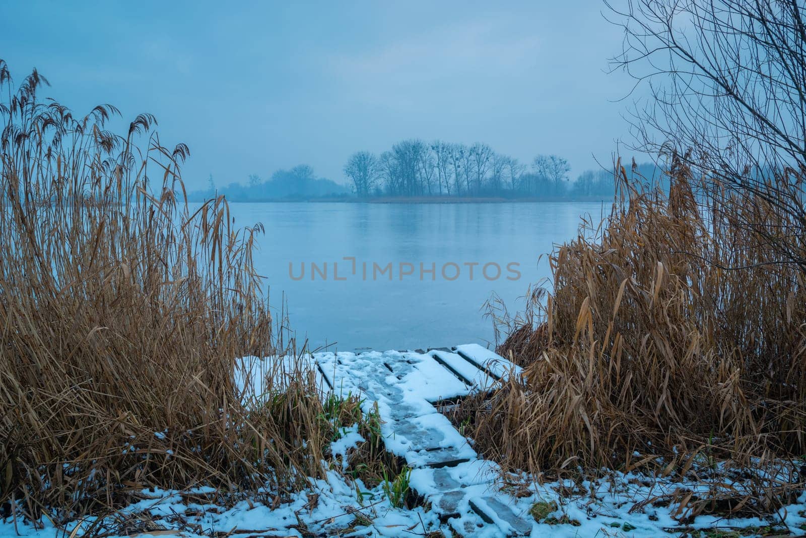 A snow-covered wooden platform in the reeds and a frozen misty lake by darekb22