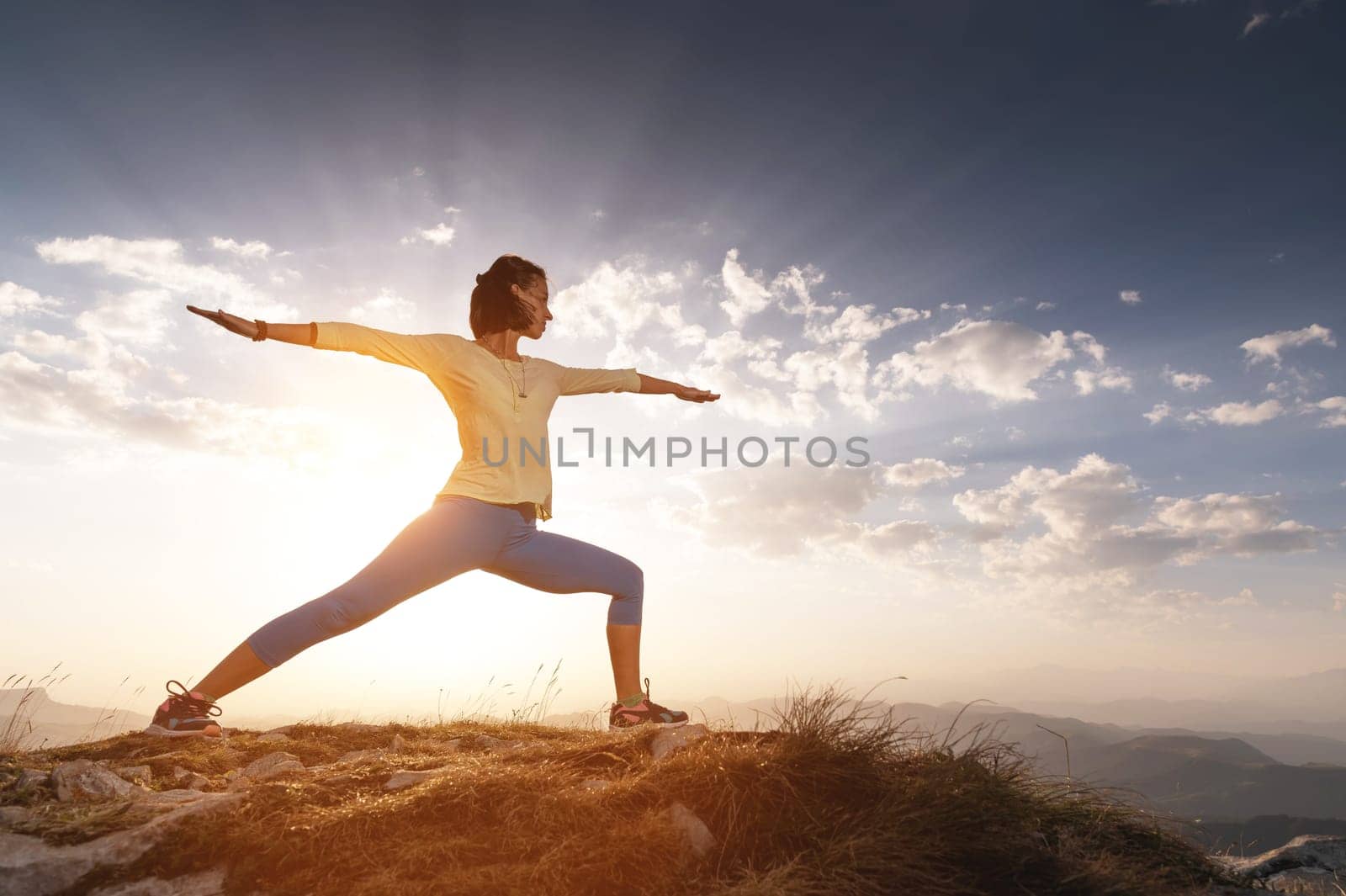 A young woman practices yoga and fitness outdoors in a beautiful mountain landscape. Morning dawn, warrior pose.