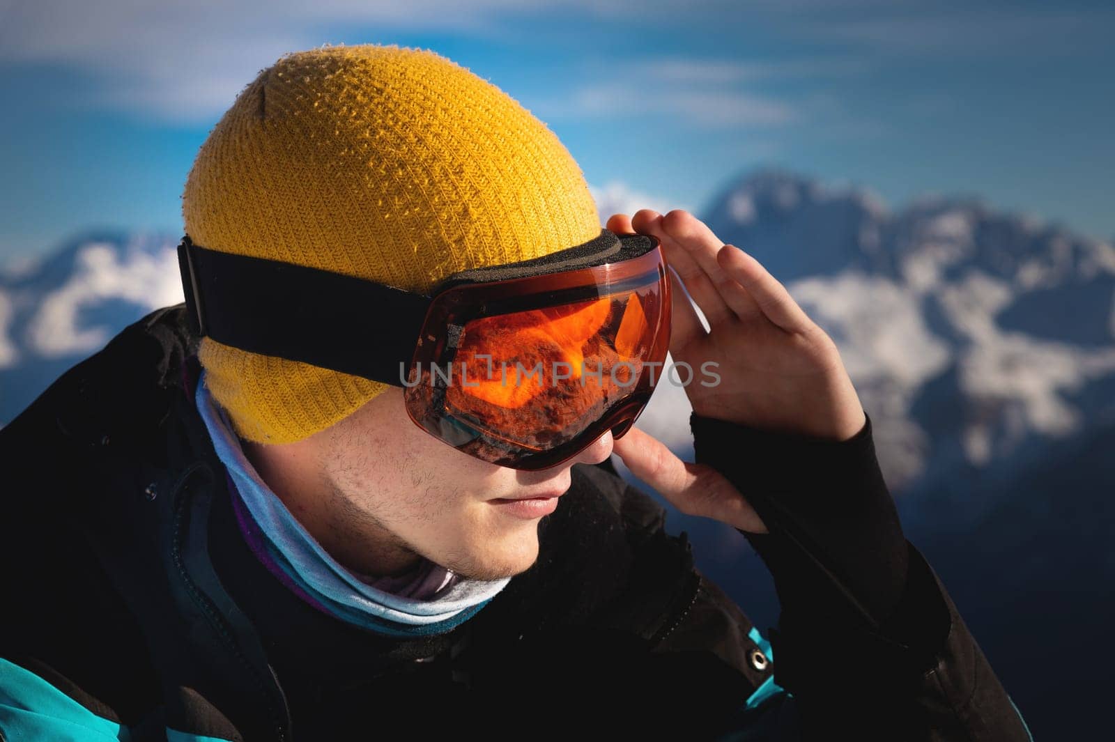 Portrait of a professional skier athlete in a blue jacket, black and orange mask looks to the side. Background with snow covered mountains of ski resort by yanik88