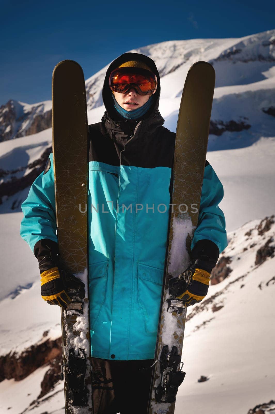 Skier confident sports man in goggles mask, spends extreme weekend in the mountains, holds skis next to him in his hands.
