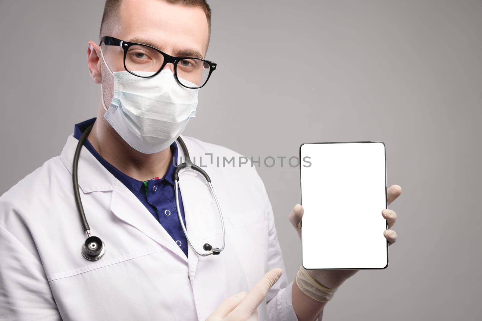 A young and handsome smart doctor wearing a surgical hygiene protective mask on a gray background and holding a tablet computer in his hand with a blank mockup screen. Idea for doctors.