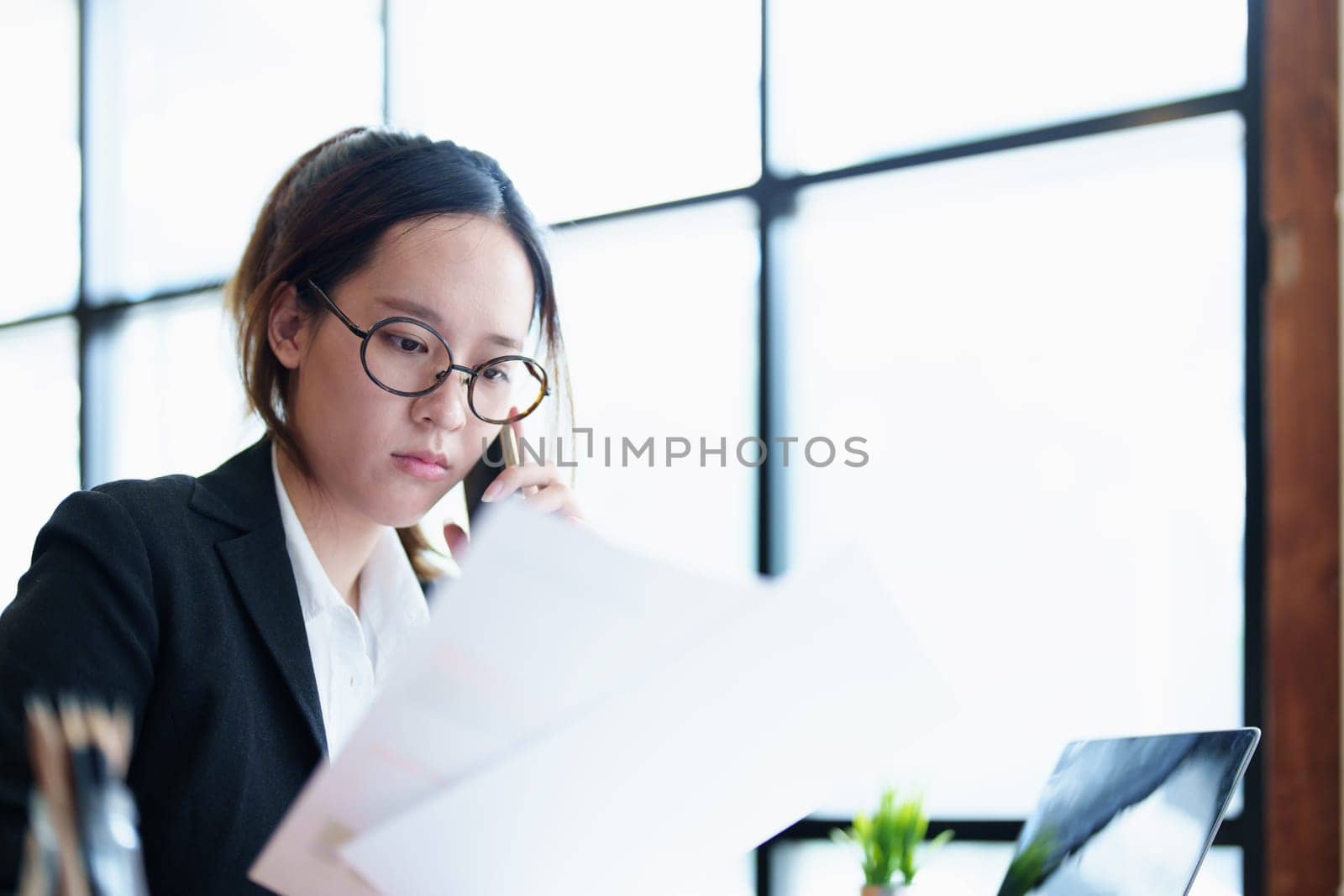 Portrait of a young Asian woman showing a serious face as she uses her phone, financial documents and computer laptop on her desk in the early morning hours by Manastrong