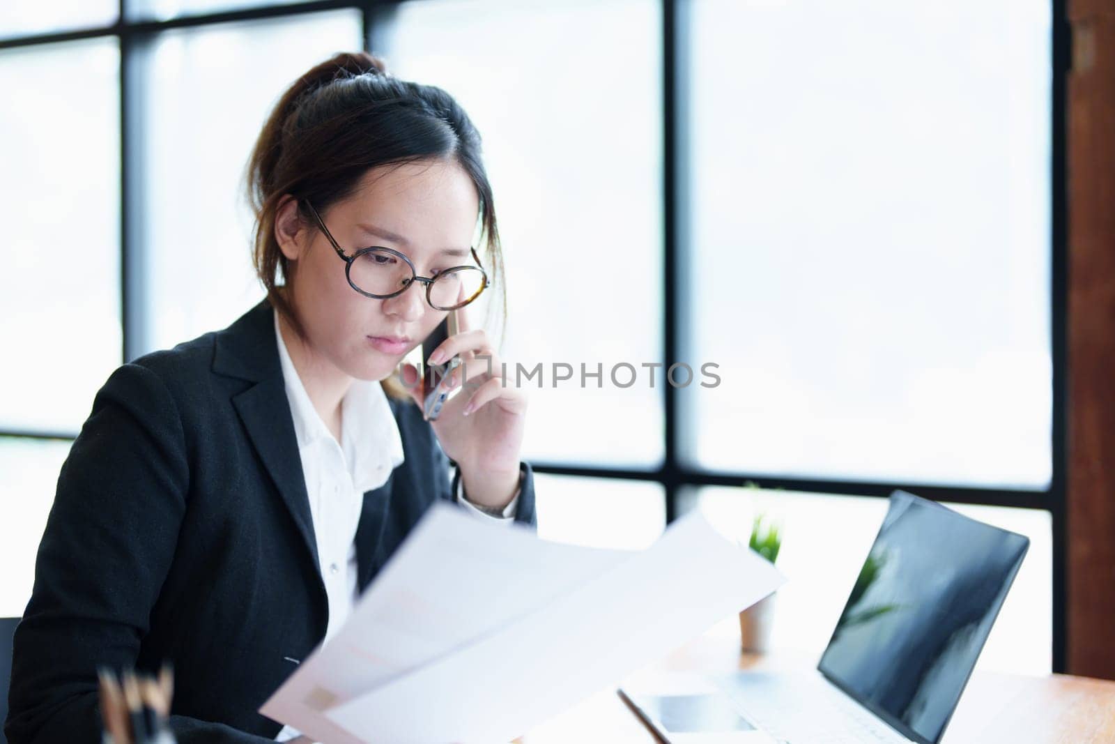 Portrait of a young Asian woman showing a serious face as she uses her phone, financial documents and computer laptop on her desk in the early morning hours.
