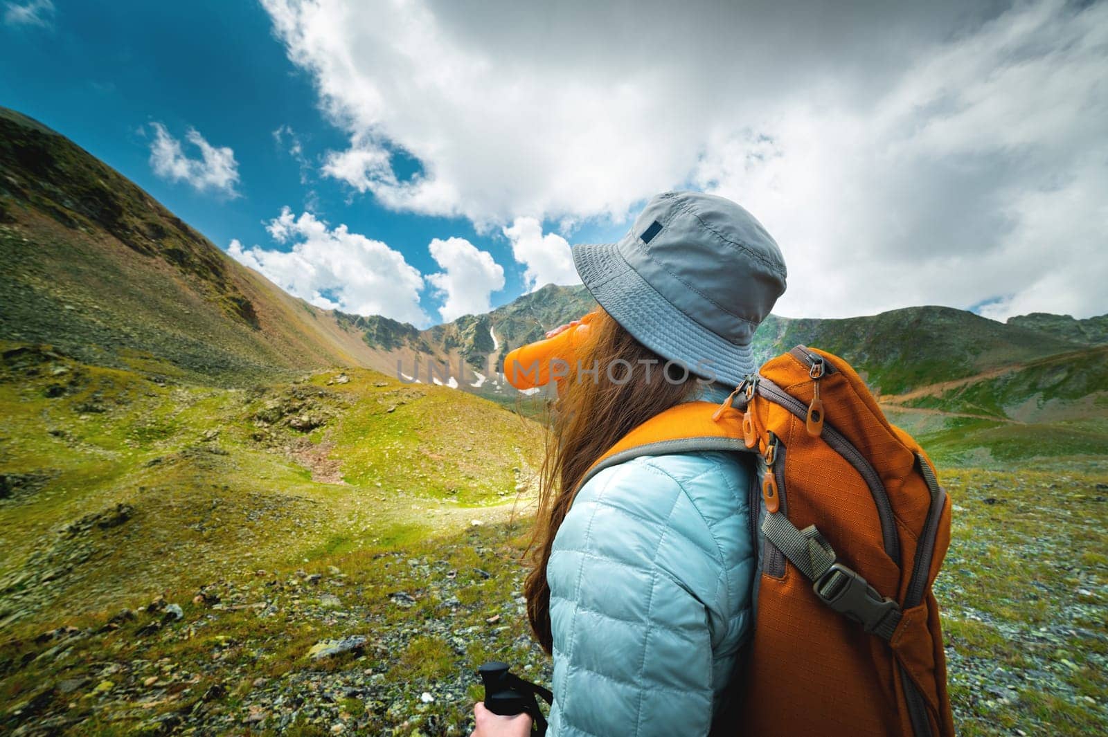A successful tourist drinks water in a green meadow near high mountains. Travel and hiking concept with backpack, side view.