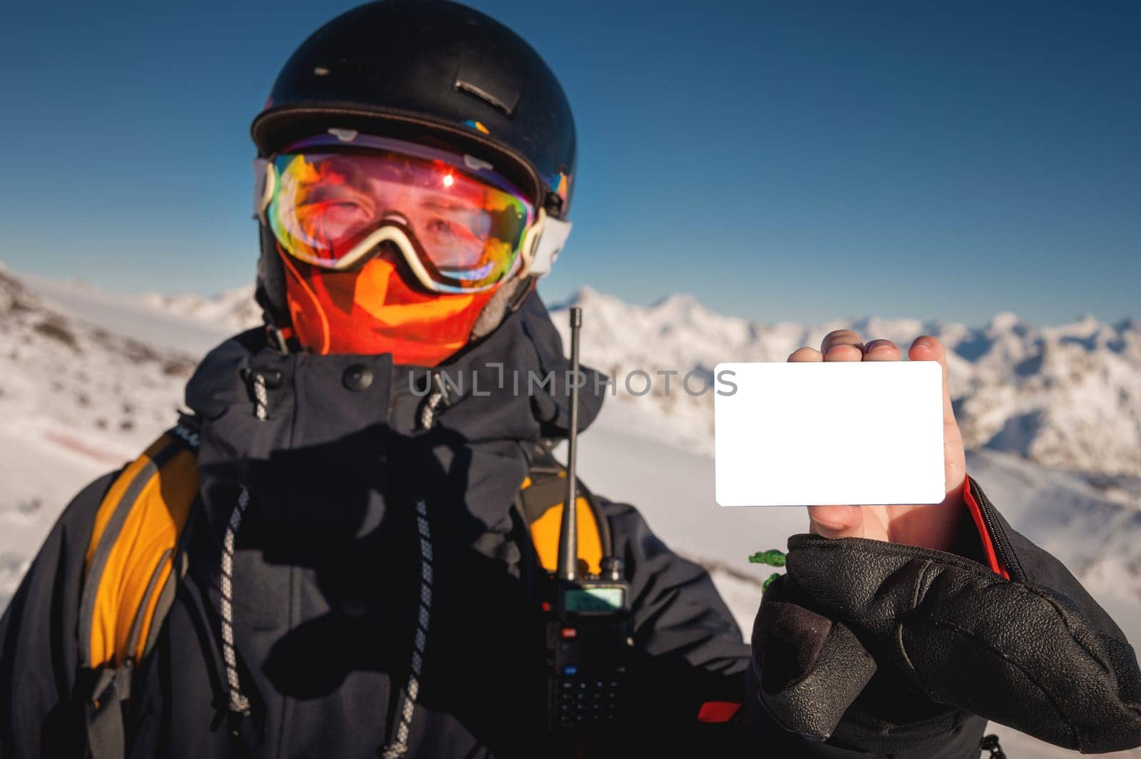 A snowboarder holds an empty lift pass with a mountain in the background. Blank ski pass in the hand of a young skier in winter gear looking at the camera. Concept illustrating ski entry fee.