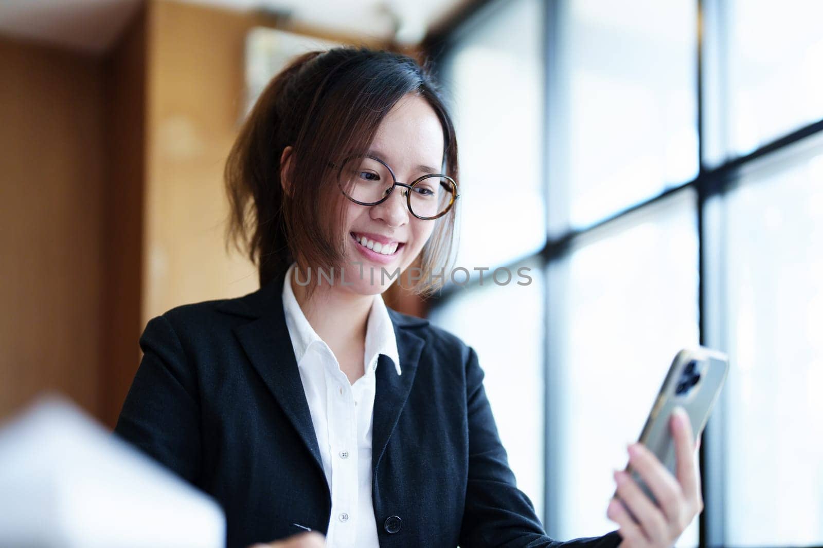 Portrait of a young Asian woman showing a smiling face as she uses her phone, computer and financial documents on her desk in the early morning hours by Manastrong