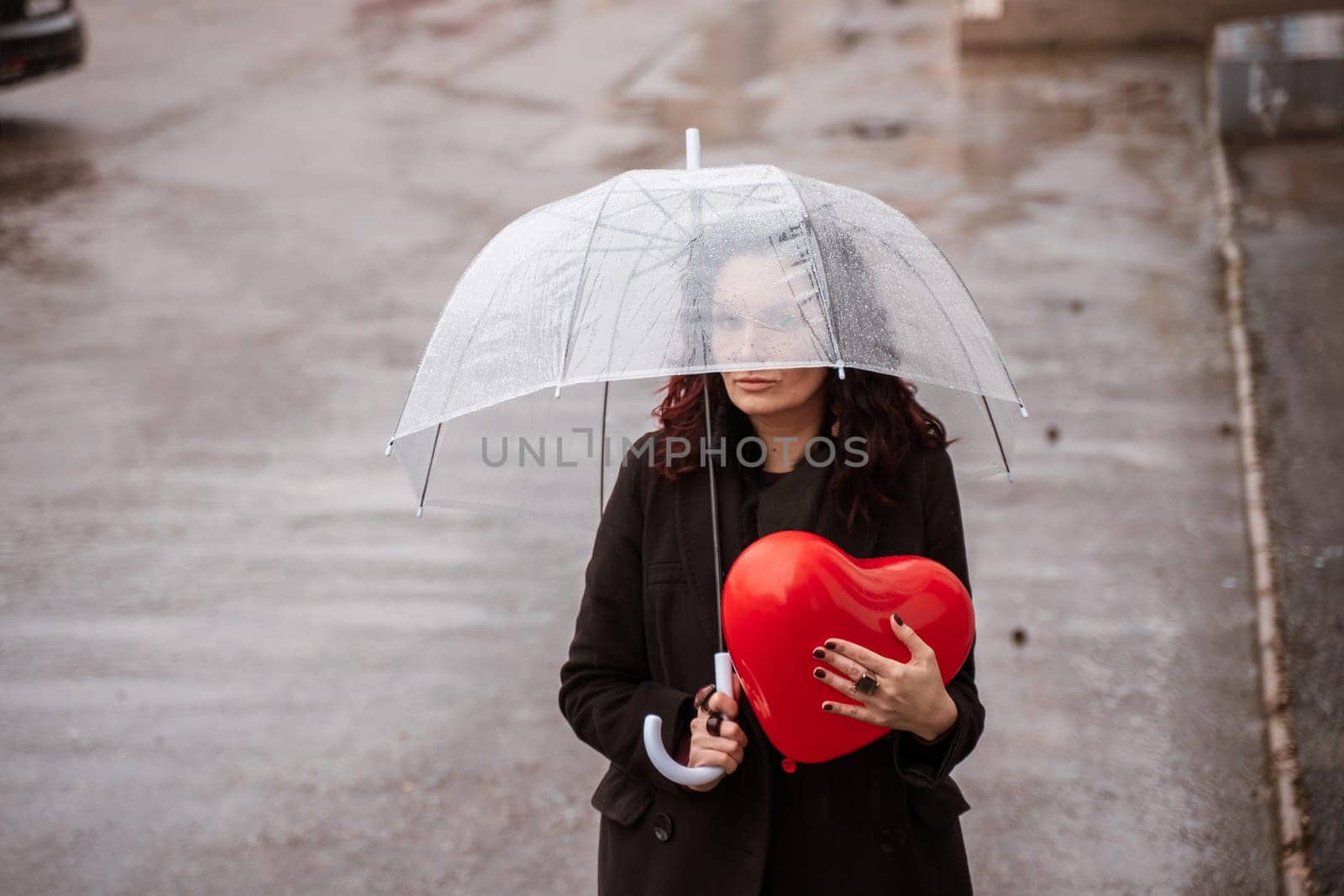 Sad middle-aged woman in a black coat and boots under a transparent umbrella. He holds a balloon in the form of a heart in his hands. Rain in the city, outdoor scene, water drops on the umbrella. The concept of loneliness, unhappy love