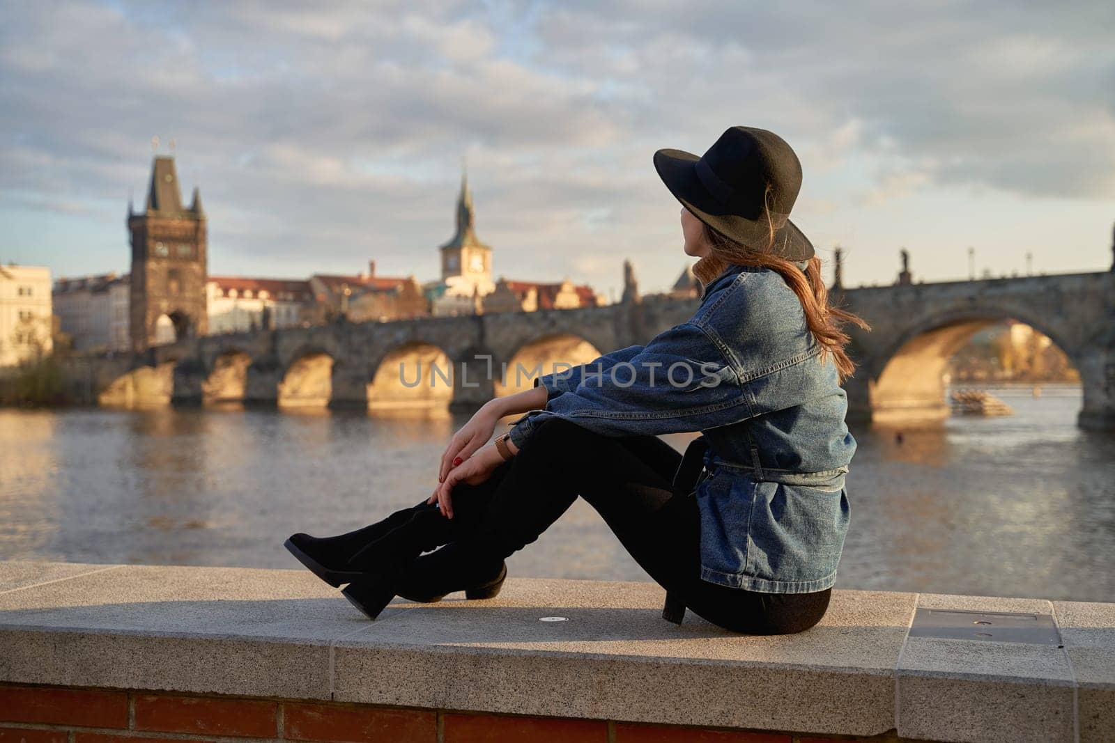 Stylish beautiful young woman wearing black hat sitting on Vltava river shore in Prague with Charles Bridge on background. Elegant retro lady fine art portrait. by berezko