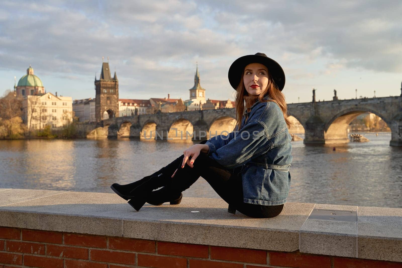 Stylish beautiful young woman wearing black hat sitting on Vltava river shore in Prague with Charles Bridge on background. Elegant retro lady fine art portrait. by berezko