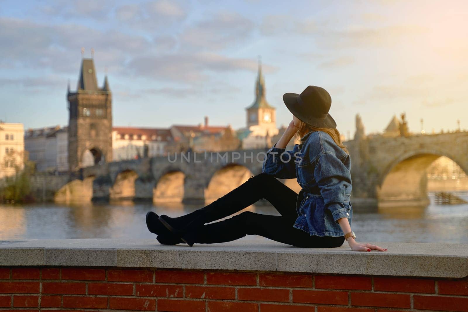 Stylish beautiful young woman wearing black hat sitting on Vltava river shore in Prague with Charles Bridge on background. Focus on foreground. by berezko