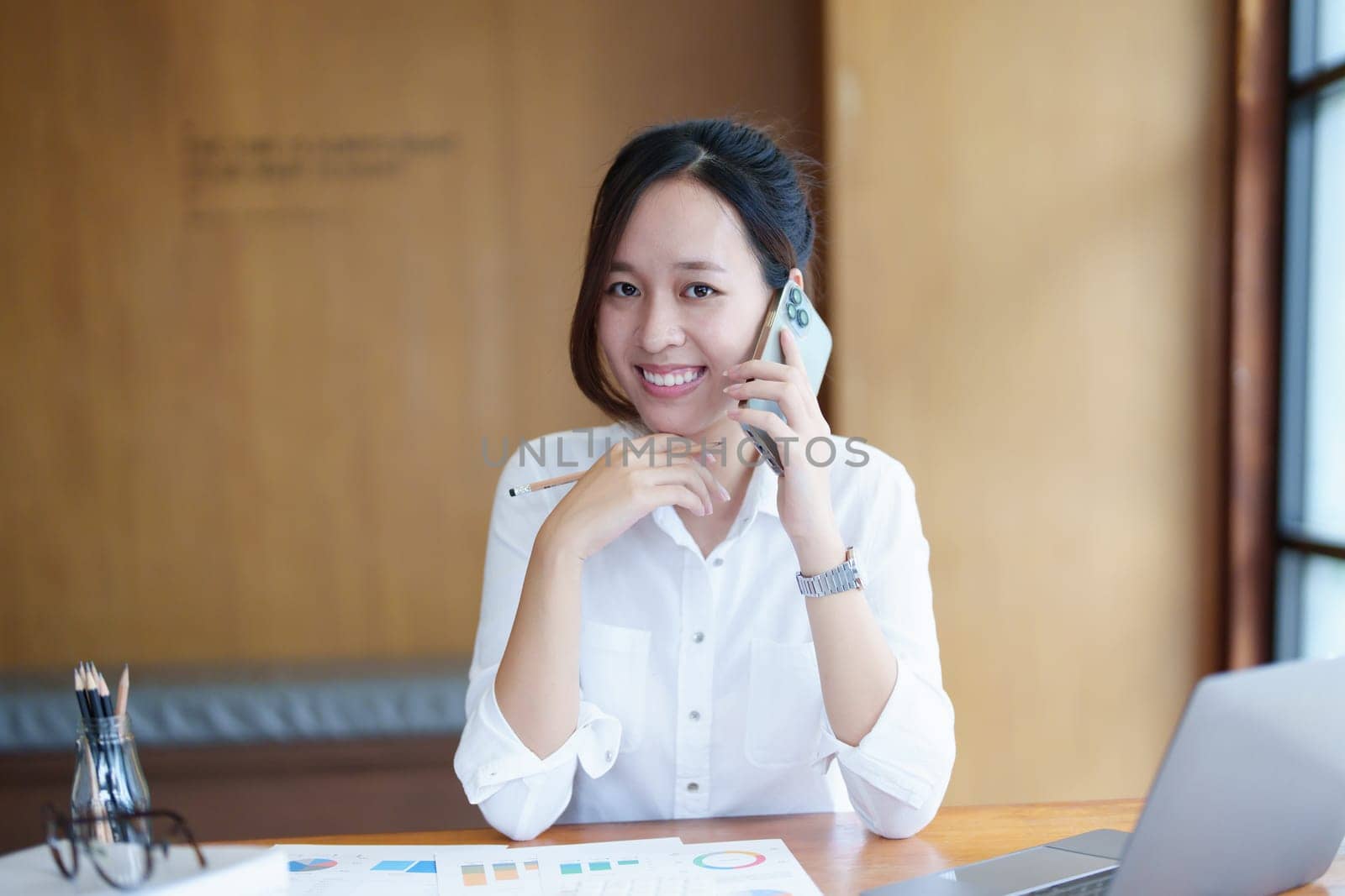 Portrait of a young Asian woman showing a smiling face as she uses her phone, computer and financial documents on her desk in the early morning hours.