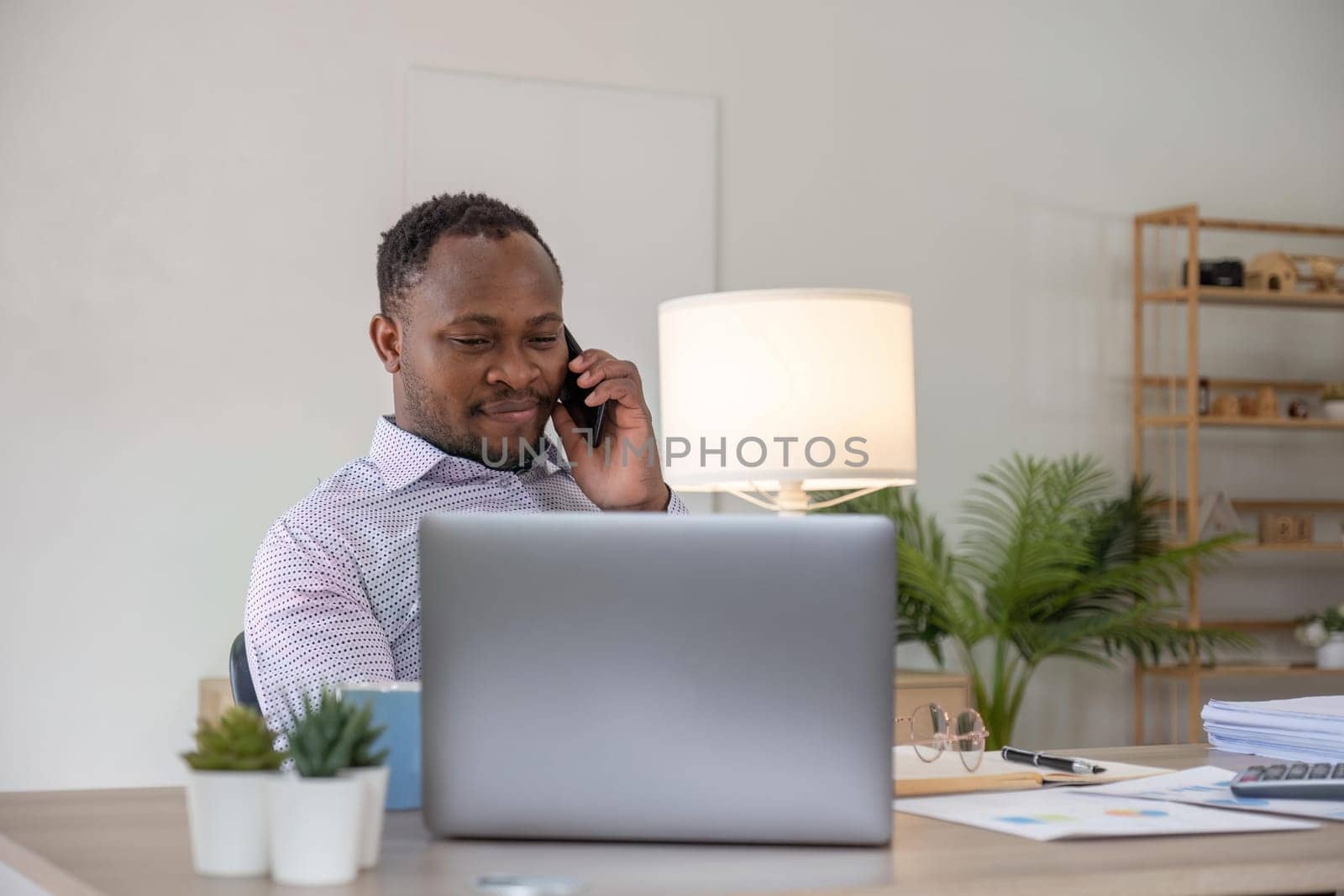 Happy black businessman talking on cellphone having phone conversation sitting at workplace working on laptop computer in office..