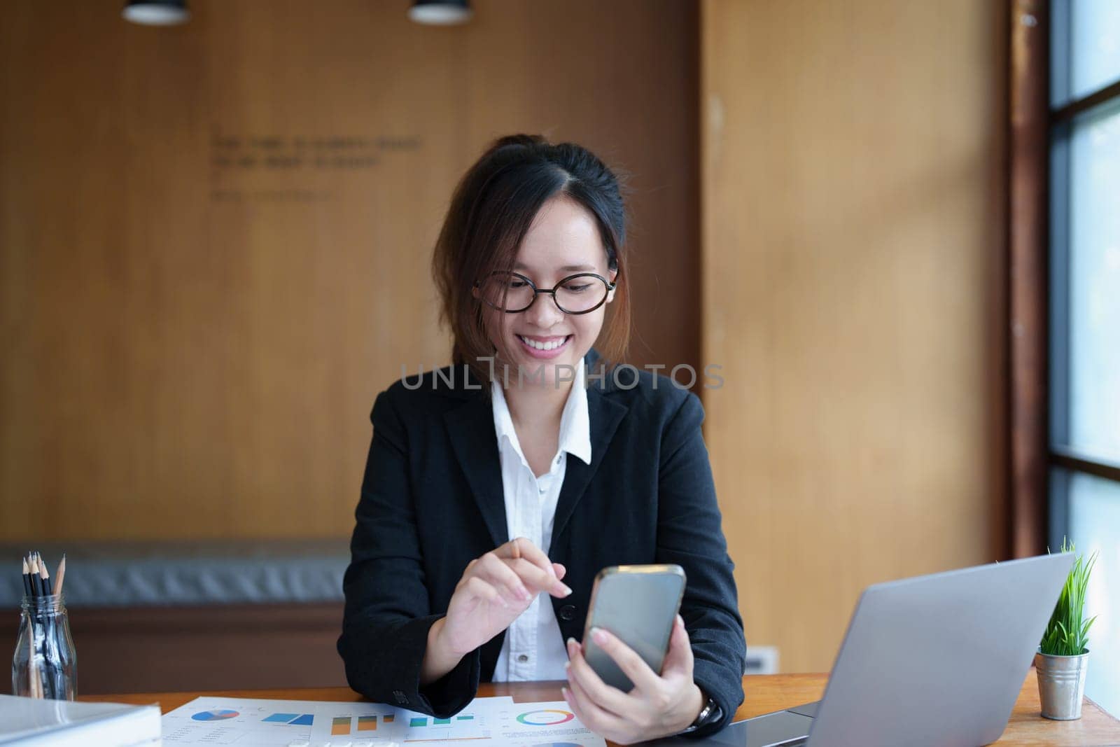 Portrait of a young Asian woman showing a smiling face as she uses her phone, computer and financial documents on her desk in the early morning hours by Manastrong