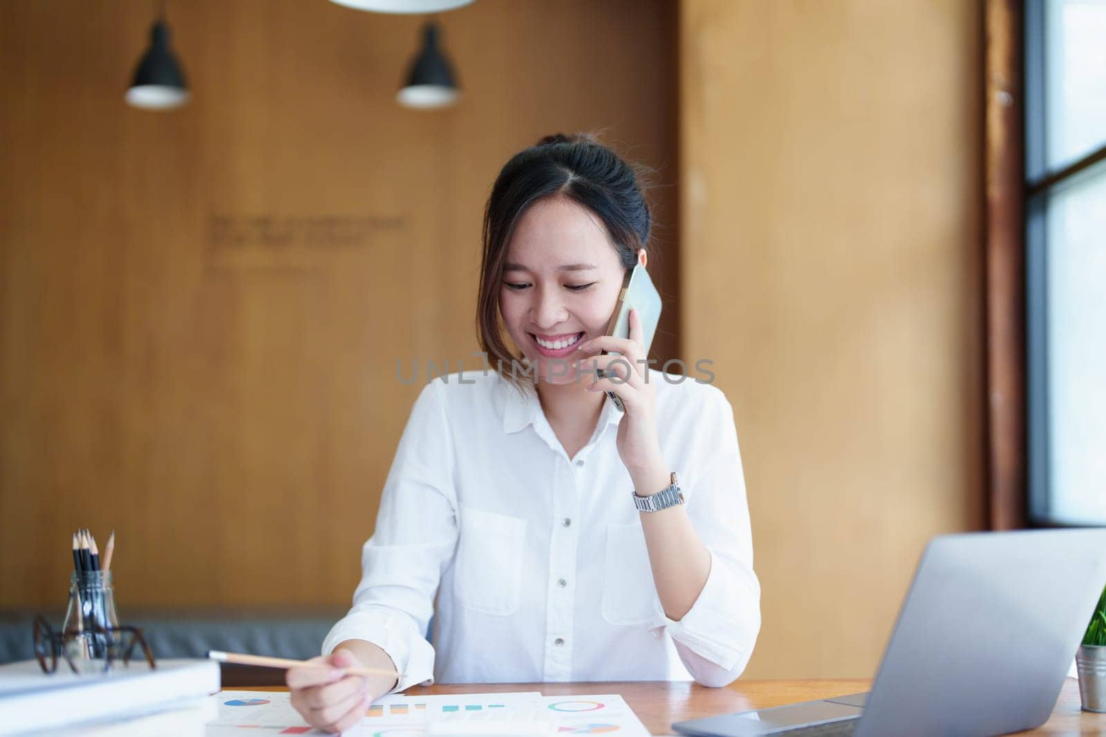 Portrait of a young Asian woman showing a smiling face as she uses her phone, computer and financial documents on her desk in the early morning hours by Manastrong