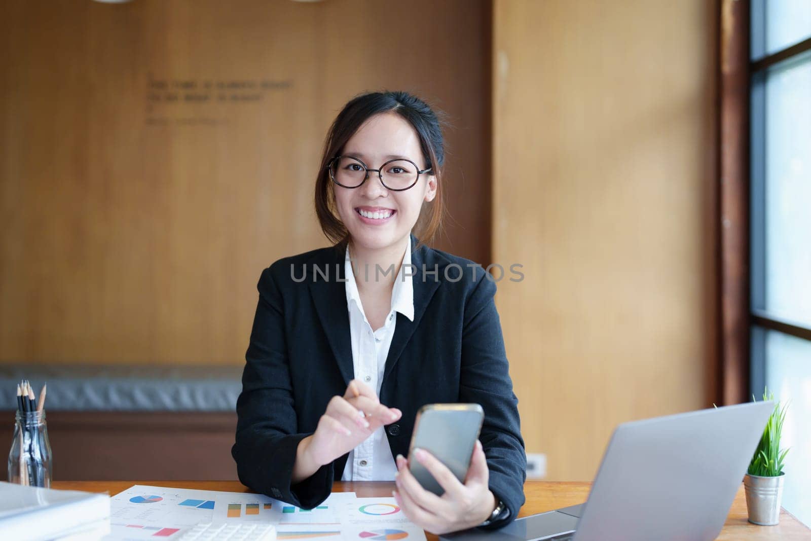 Portrait of a young Asian woman showing a smiling face as she uses her phone, computer and financial documents on her desk in the early morning hours.