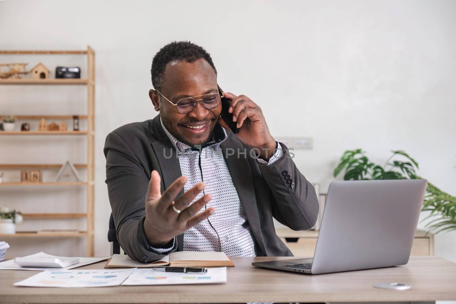 Happy black businessman talking on cellphone having phone conversation sitting at workplace working on laptop computer in office..
