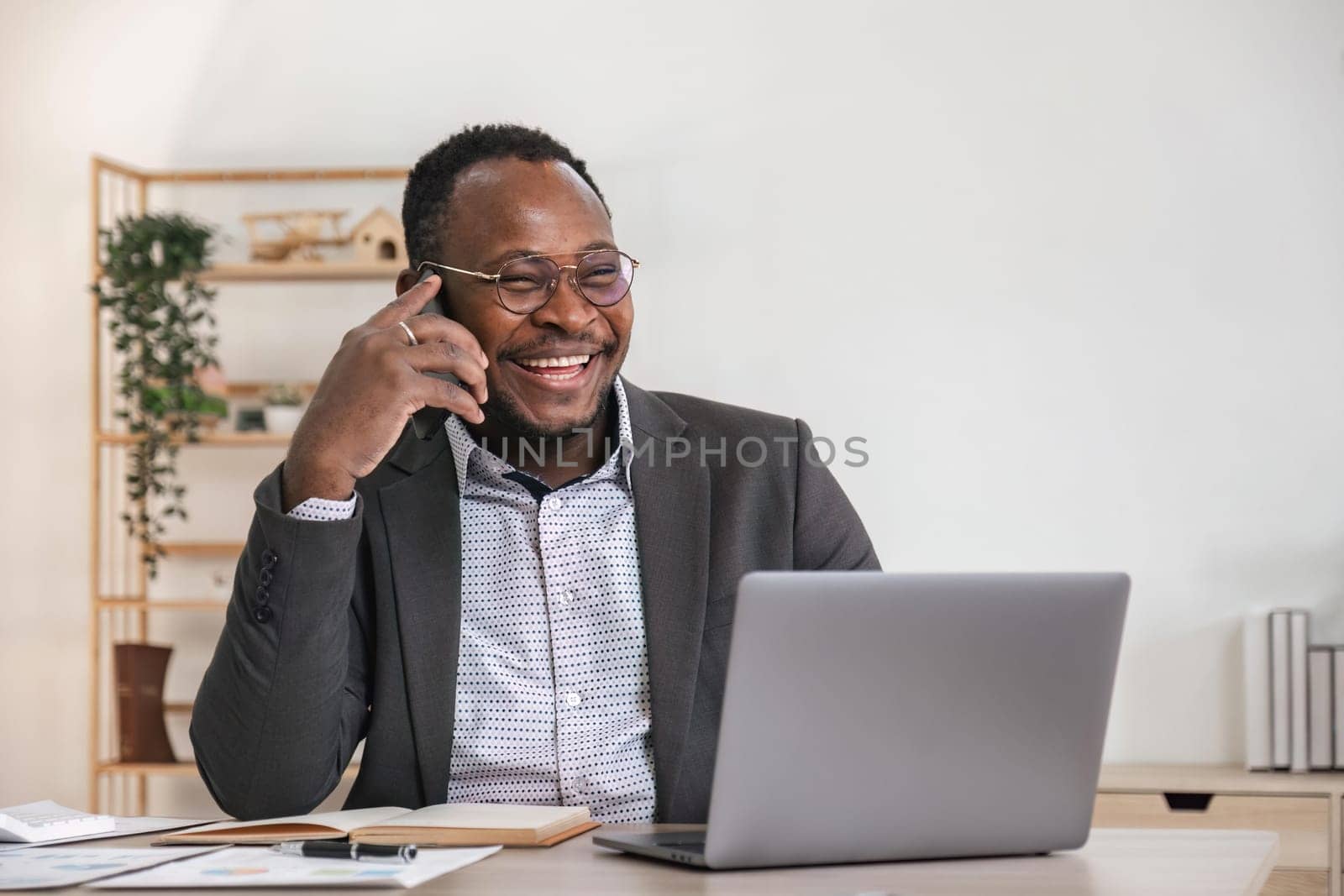 Happy black businessman talking on cellphone having phone conversation sitting at workplace working on laptop computer in office. by nateemee