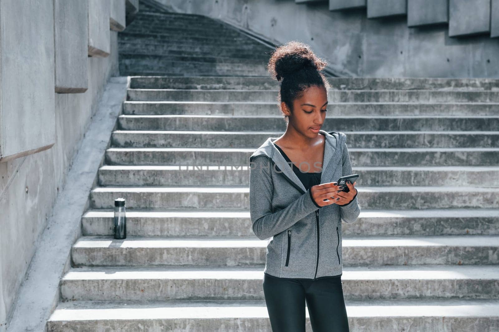 Uses phone. Young african american woman in sportive clothes have workout outdoors at daytime.