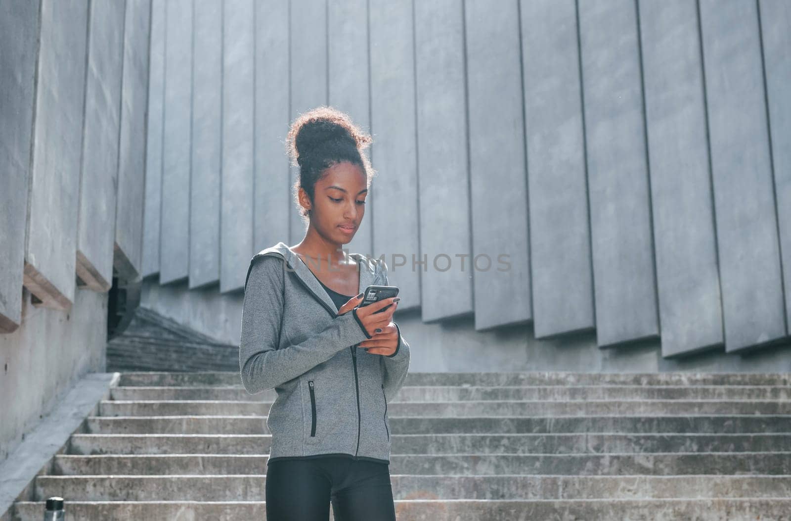 Uses phone. Young african american woman in sportive clothes have workout outdoors at daytime.