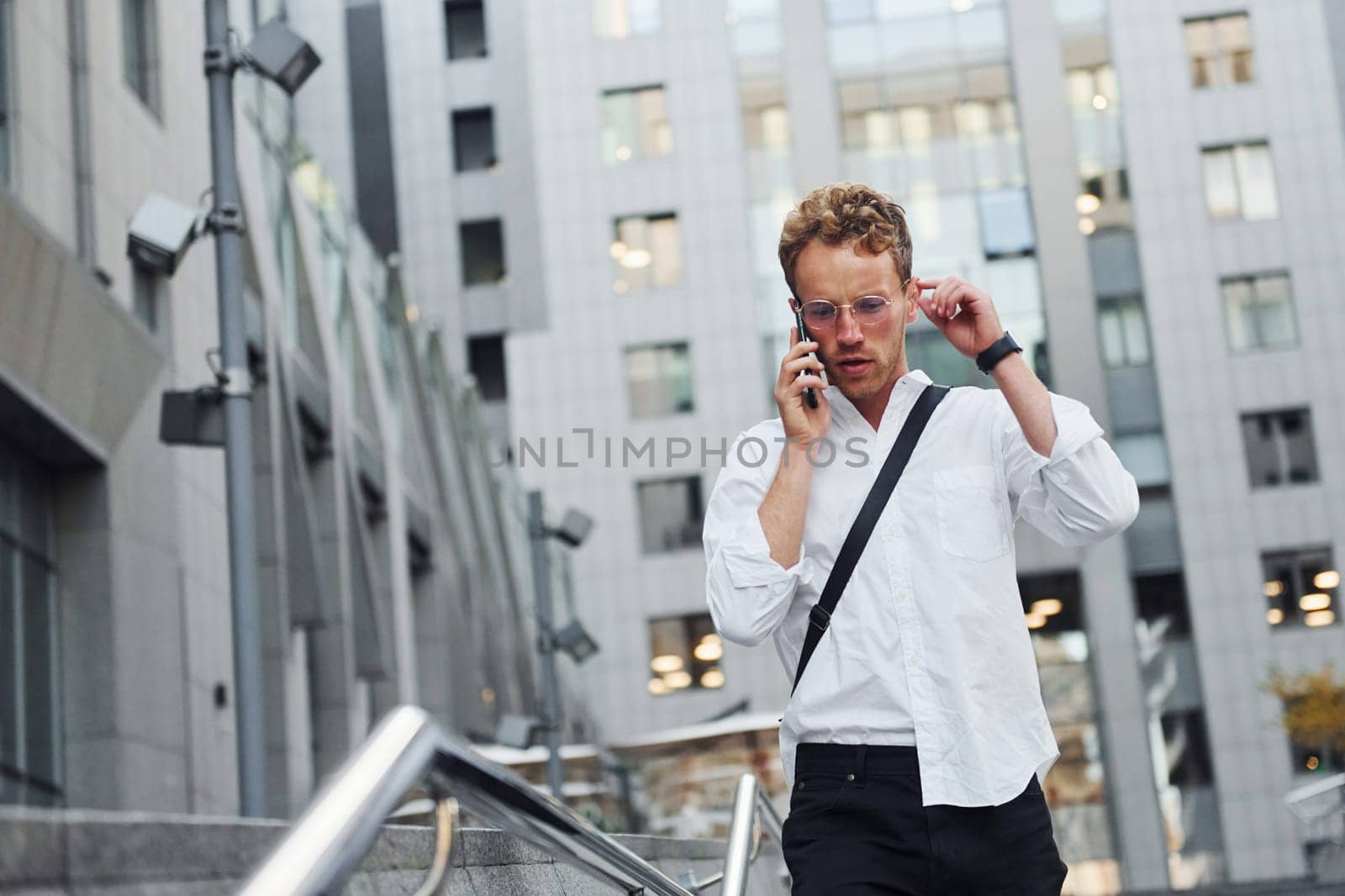 Have conversation by the phone. Young elegant man in good clothes is outdoors in the city at daytime.