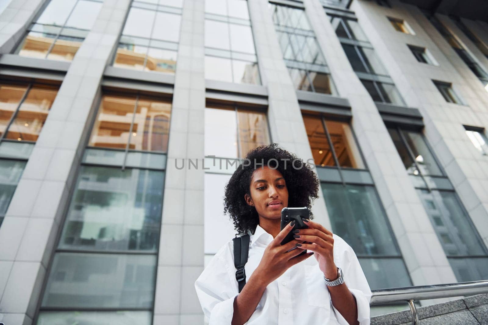 Uses phone. African american woman in good clothes is outdoors in the city at daytime.