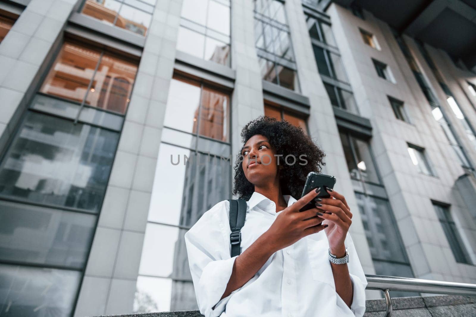 Uses phone. African american woman in good clothes is outdoors in the city at daytime.