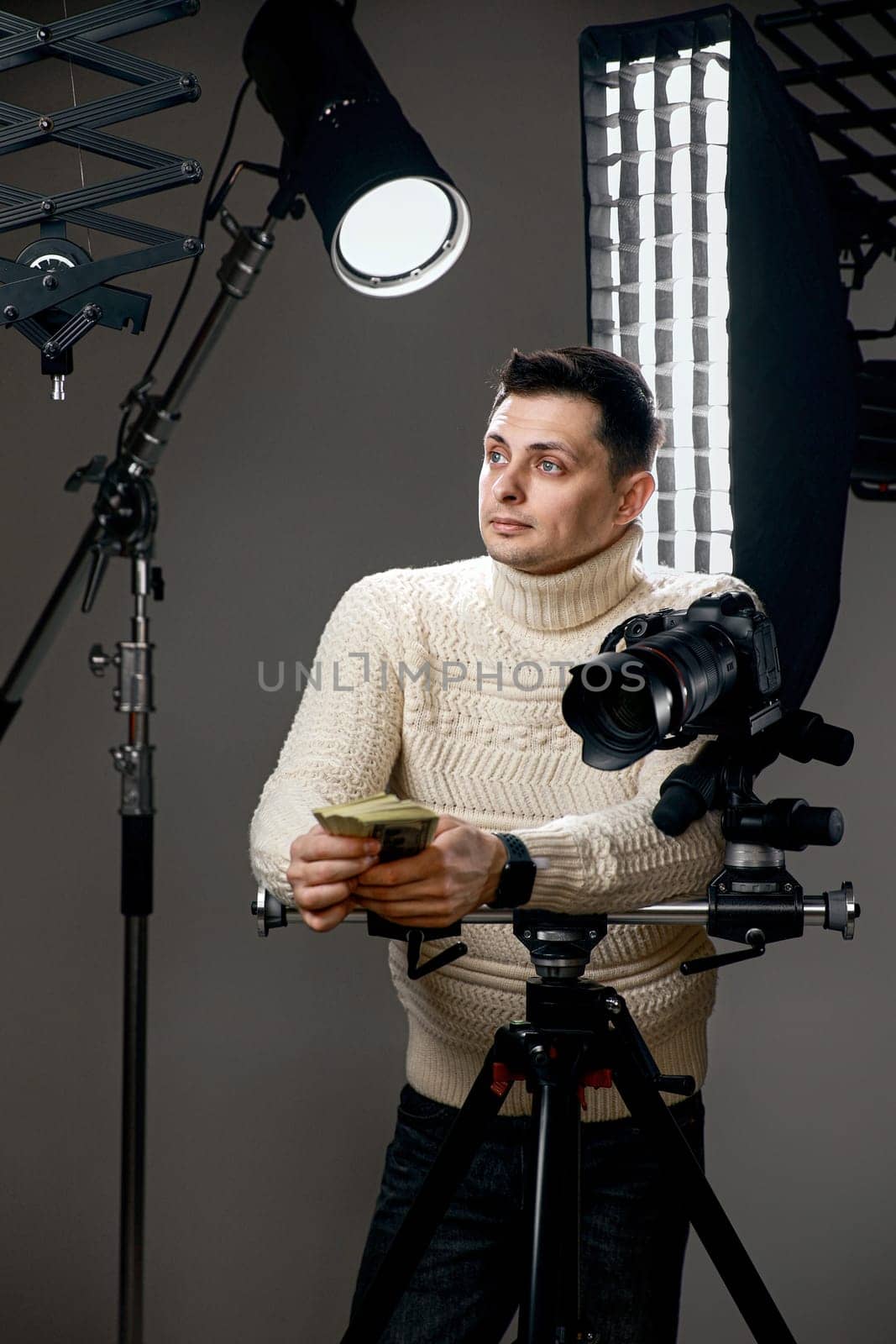 Professional handsome photographer with digital camera on tripod holding money on gray background with lighting equipment