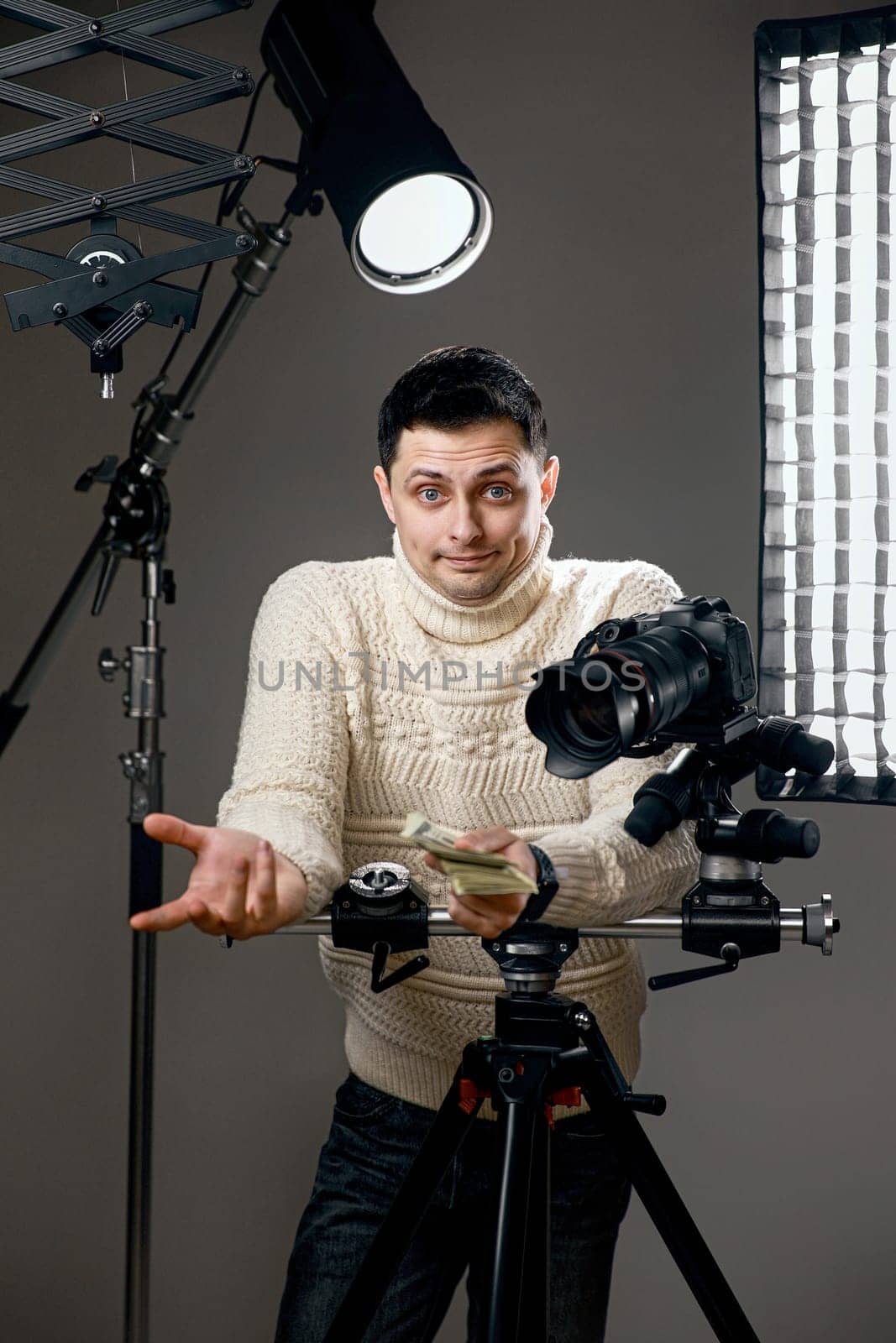 surprised professional photographer with digital camera on tripod holding hundred dollar bills on gray background with lighting equipment