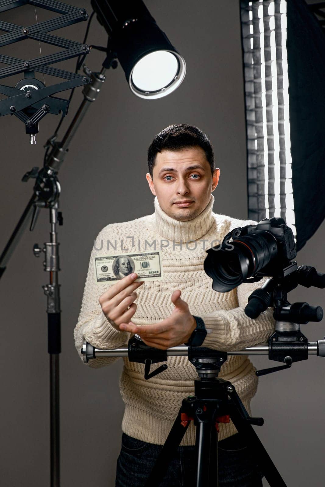 Professional handsome photographer with digital camera on tripod showing hundred dollar bill on gray background with lighting equipment