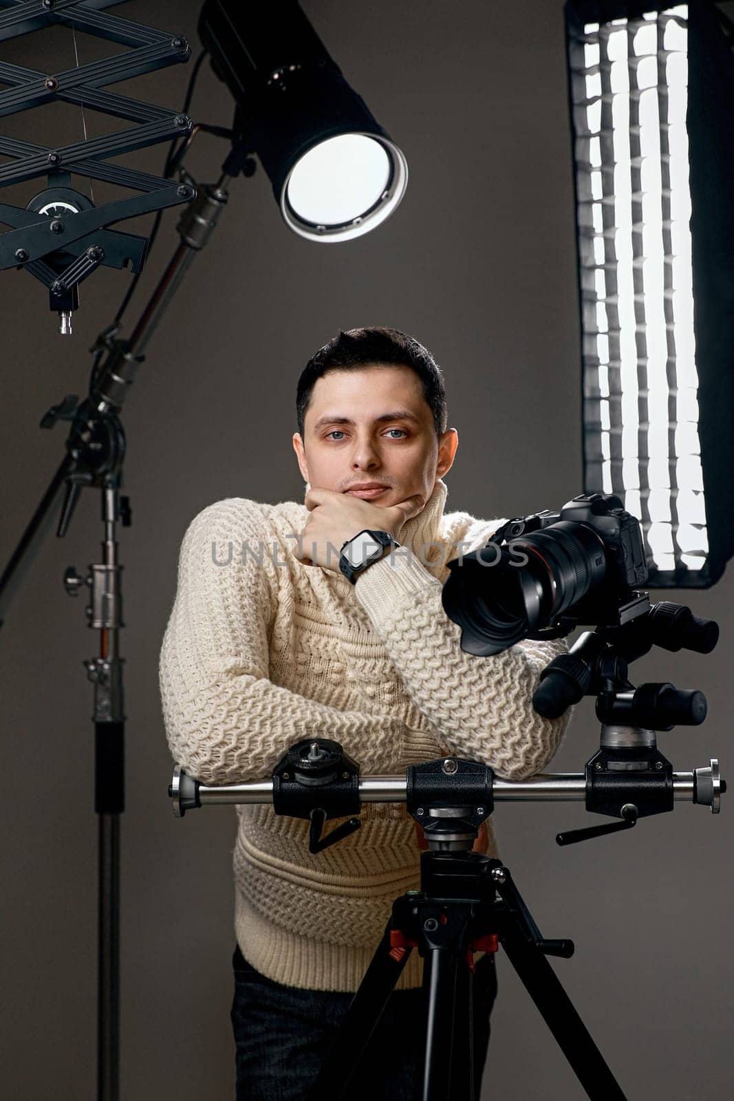 Positive professional photographer with digital camera on tripod on gray background with lighting equipment
