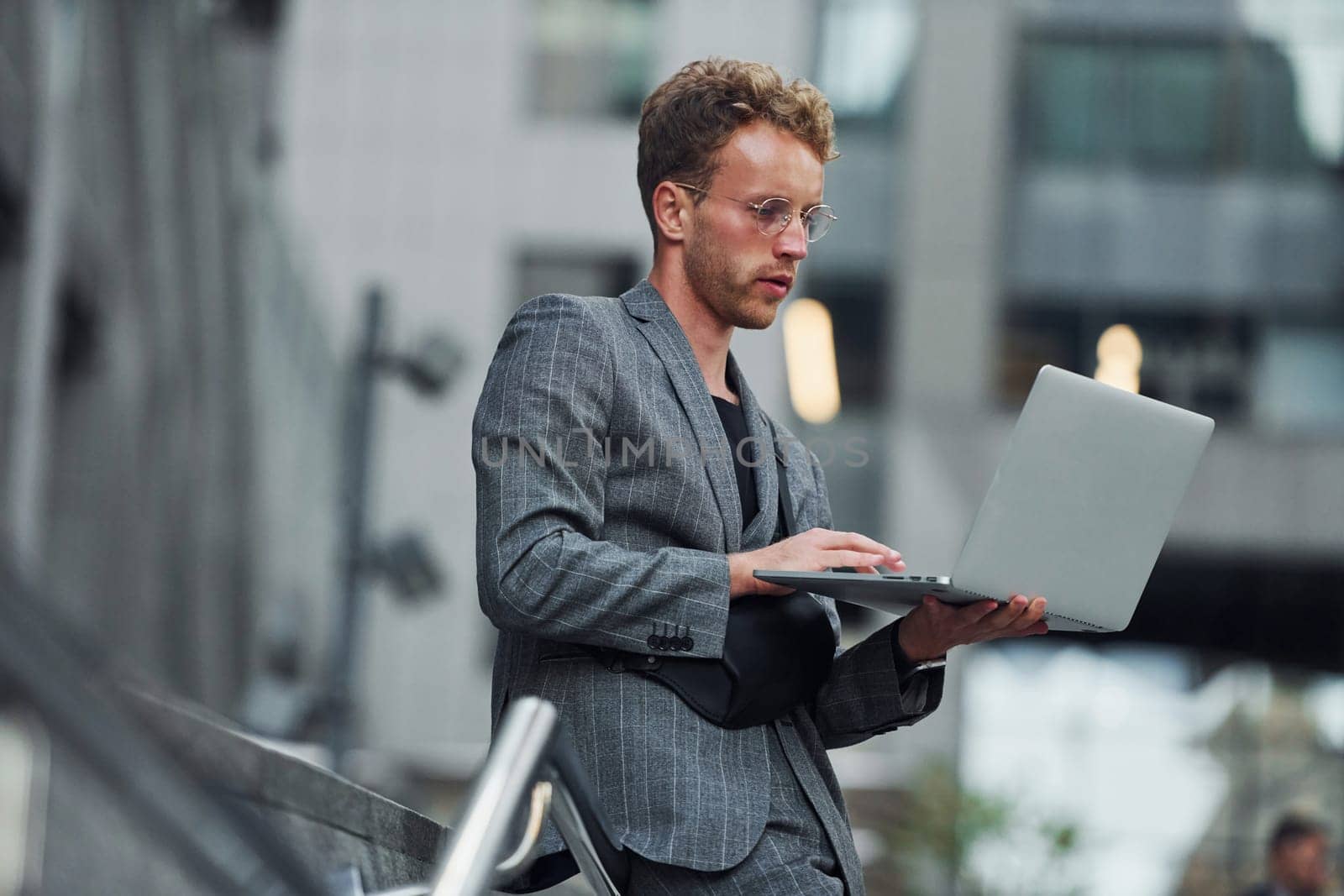 Laptop in hands. Young elegant man in good clothes is outdoors in the city at daytime.