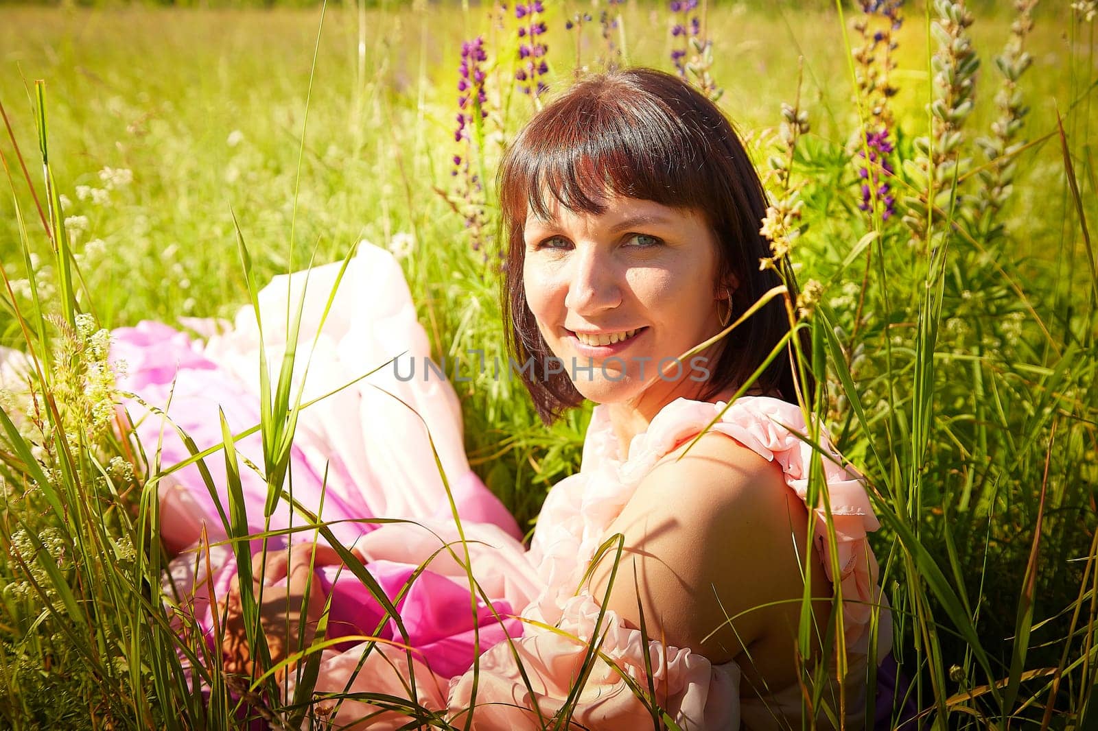 Beautiful girl in lush pink ball gown in green field during blooming of flowers and blue sky on background. Model posing on nature landscape as princess from fary tale