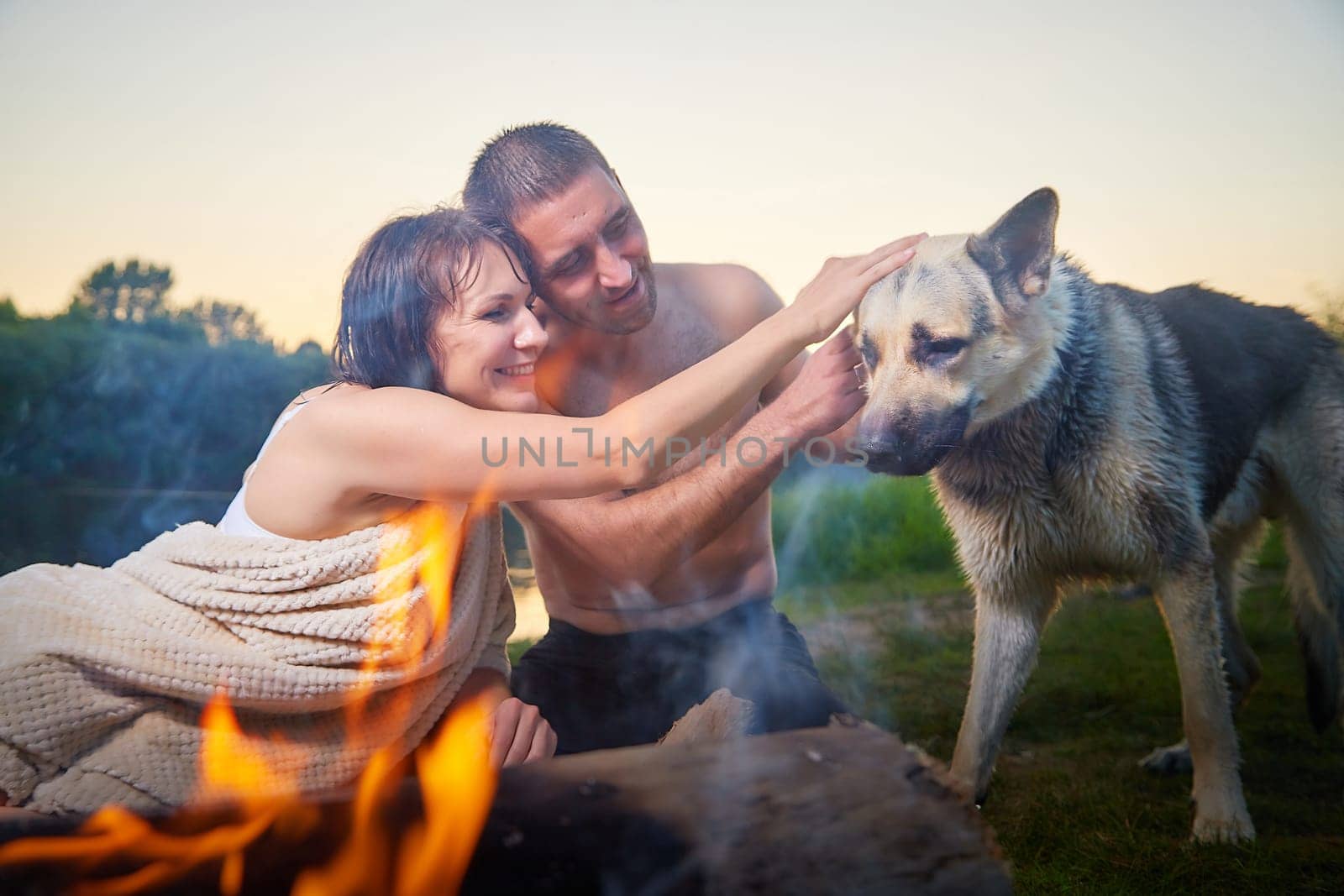 Happy wet couple relaxin, having fun and hugs with big dog near fire in camping on nature in summer sunny evening in sunset. Family or lovers have date and rest outdoor. Concept of love