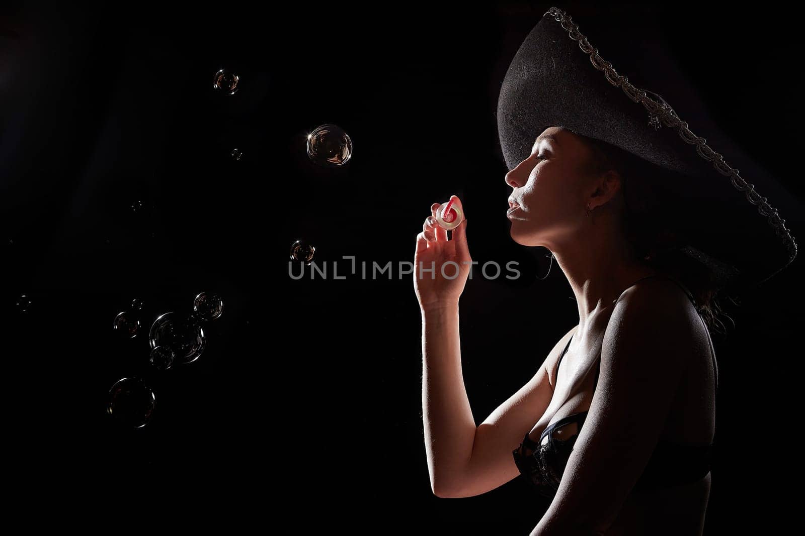 Silhouette of a beautiful young girl with soap bubbles in black underwear and a Mexican hat on dark background. Exotic model posing naked in the studio. Body portrait of sexy girl in usial photoshoot by keleny