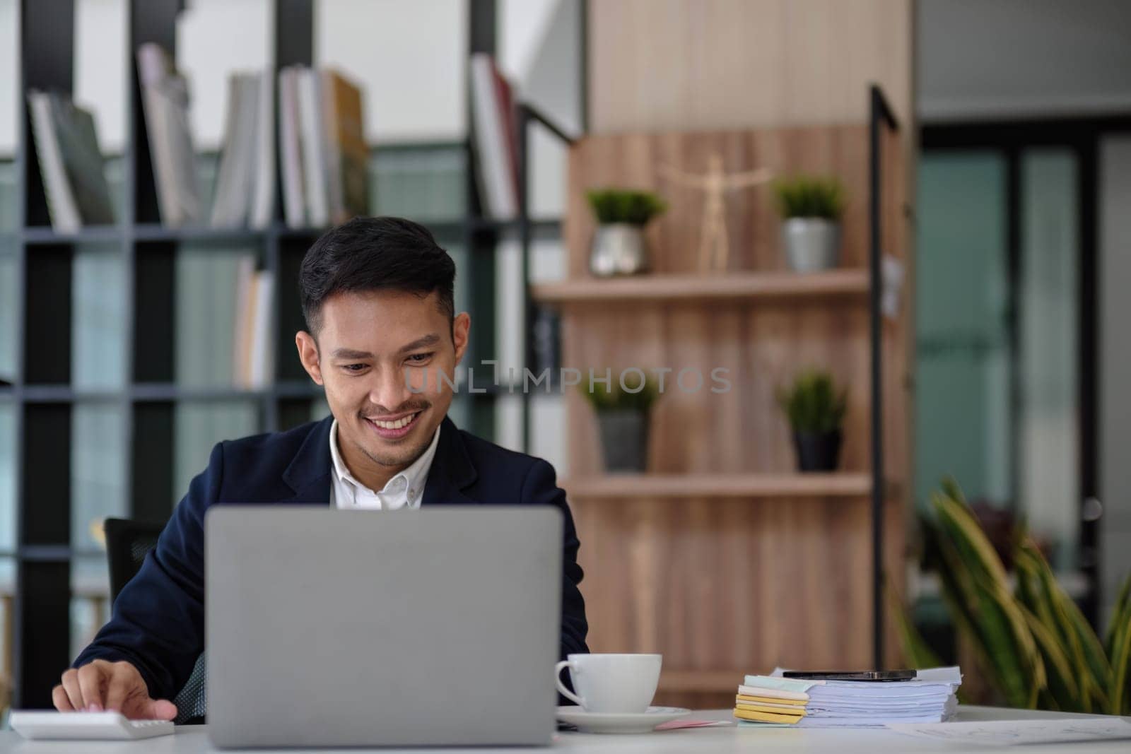 Asian business man wears a black suit and sitting in office and typing to keyboard on laptop and smiling confidently. by wichayada