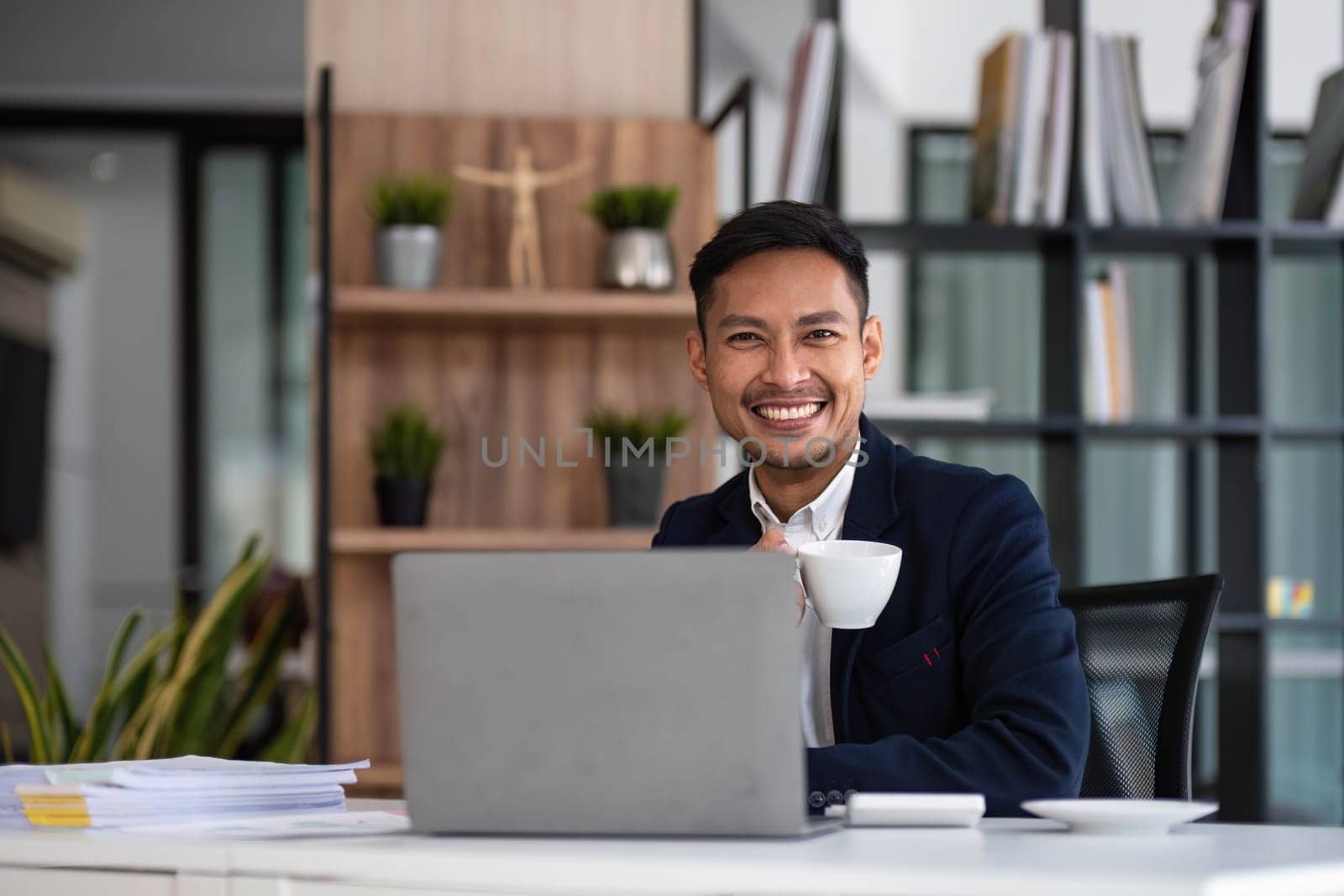 Asian business man wears a black suit and sitting in office and typing to keyboard on laptop and smiling confidently...