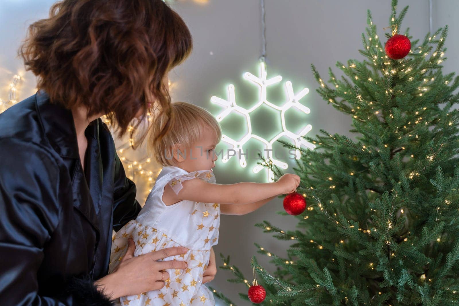 A mother with a 2-year-old daughter decorates the Christmas tree. Mom in a black suit, a girl in a white dress, her daughter hangs a red ball on the Christmas tree. Merry Christmas and New Year concept.