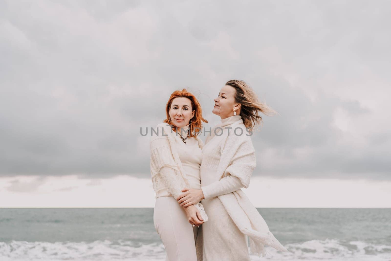 Women sea walk friendship spring. Two girlfriends, redhead and blonde, middle-aged walk along the sandy beach of the sea, dressed in white clothes. Against the backdrop of a cloudy sky and the winter sea. Weekend concept. by Matiunina