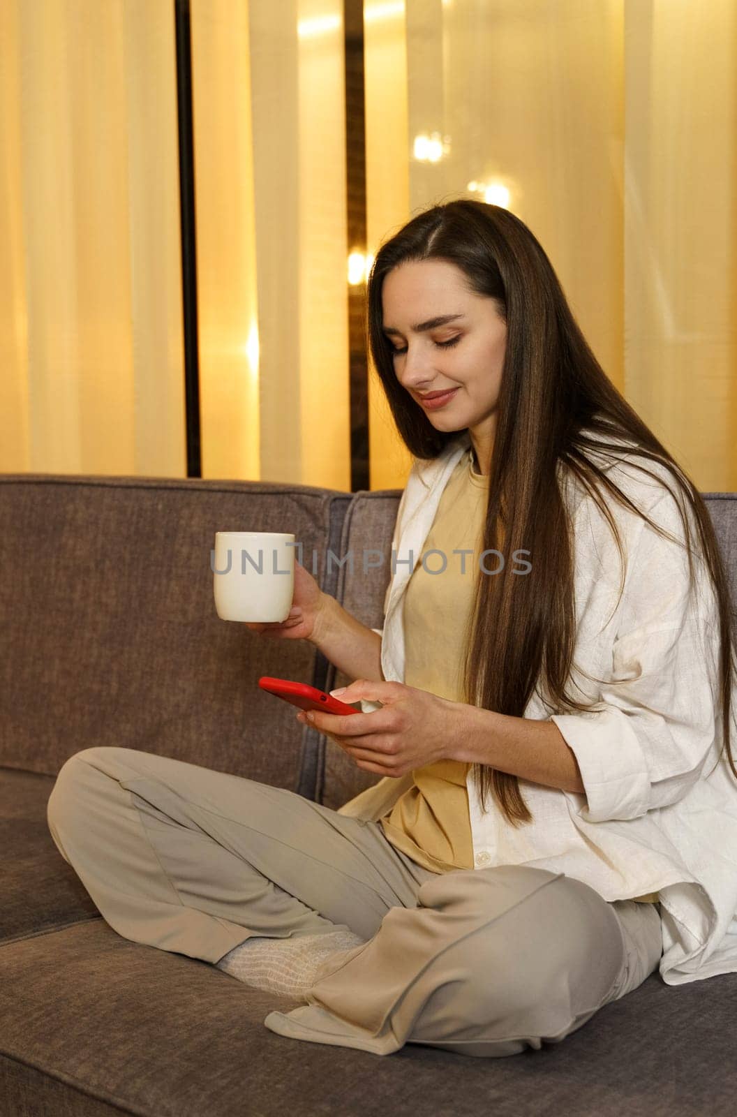 Young woman chatting with friends, surfing on the phone, enjoying her weekend at home with a cup of tea. Vertical frame.