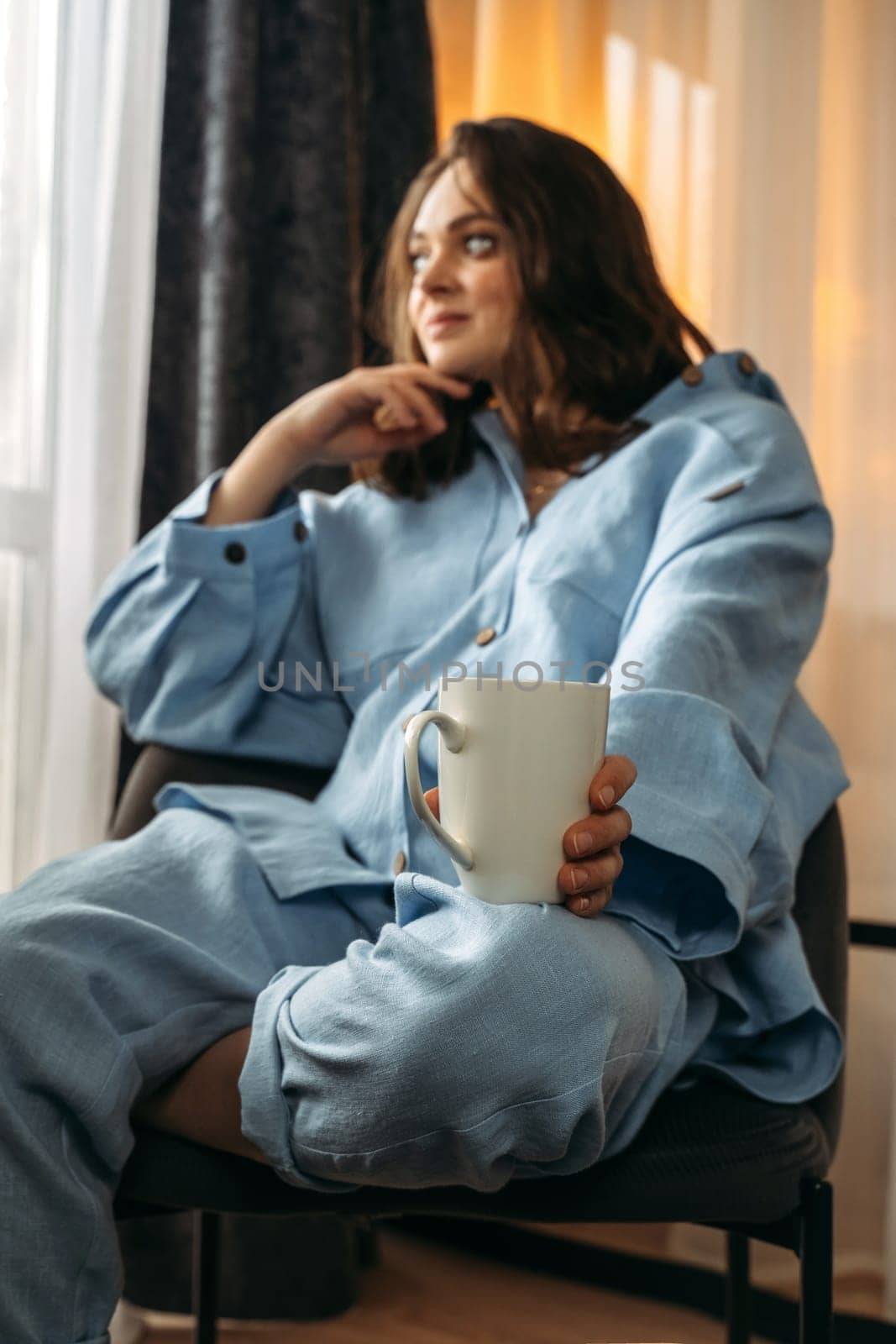 A woman has a white cup of tea or coffee on her knee. The cup is in the sharpness zone, the rest of the image is blurry. Vertical frame.