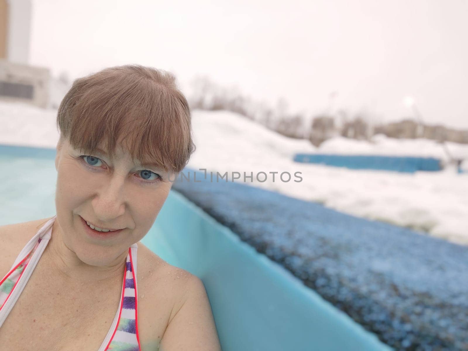 Adult mature woman takes selfie in a pool with warm hot termal mineral water in winter and white snow around. Wellness center and the concept of health care. Travel, recreation, medicine and rest