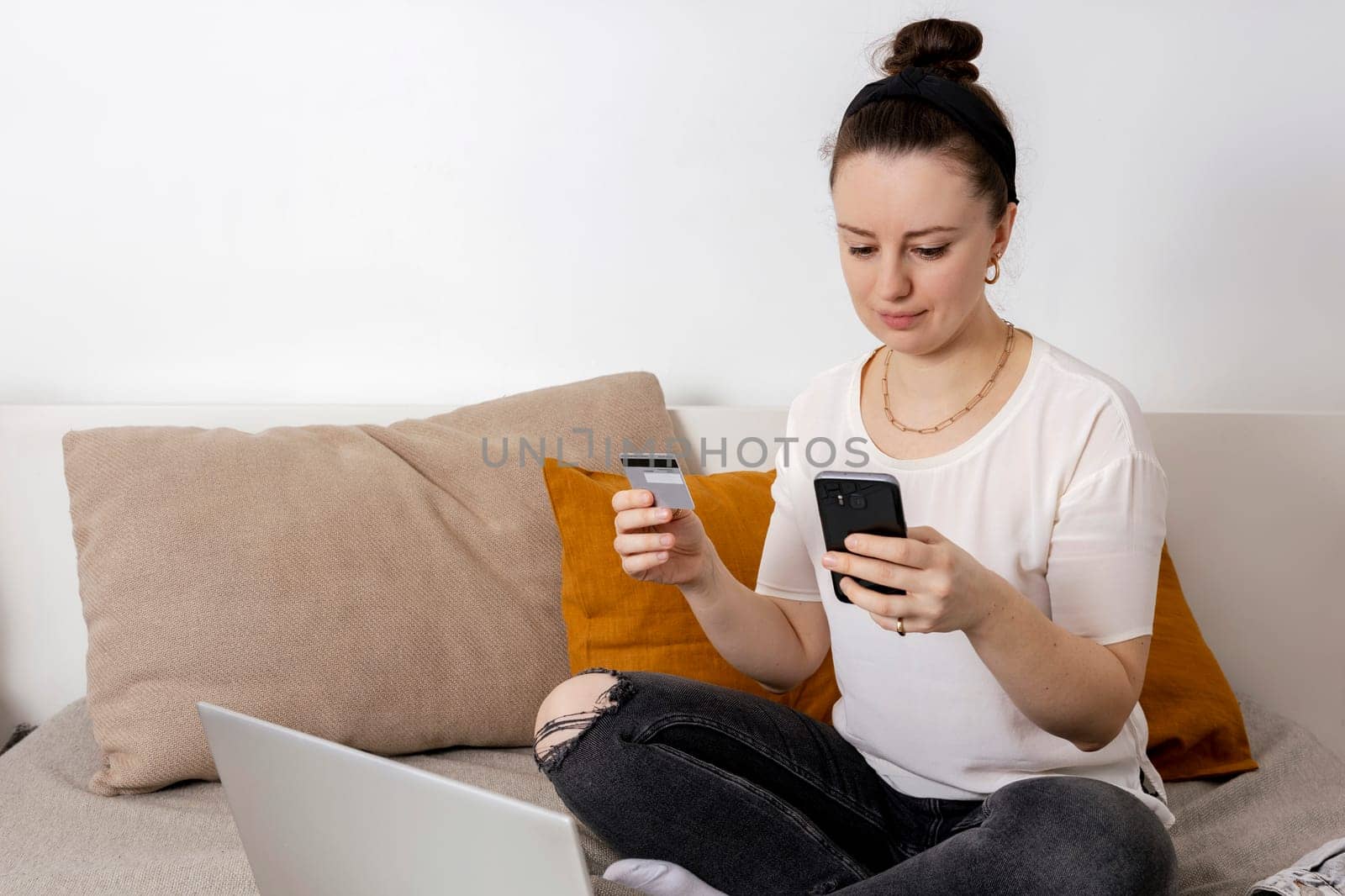 Young brunette woman smiling while surfing the web on her laptop and holding credit card in her hand trying to buy something online. Easy online shopping at home, e-commerce. Cosy interior