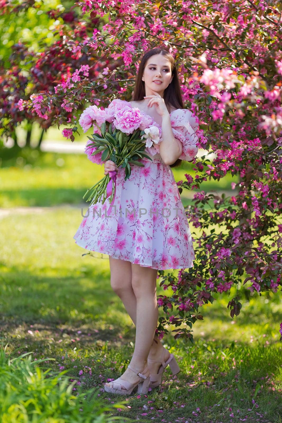 Adorable girl, stands with a large bouquet of peonies, near pink flowering trees in the garden. Copy space. Vertical