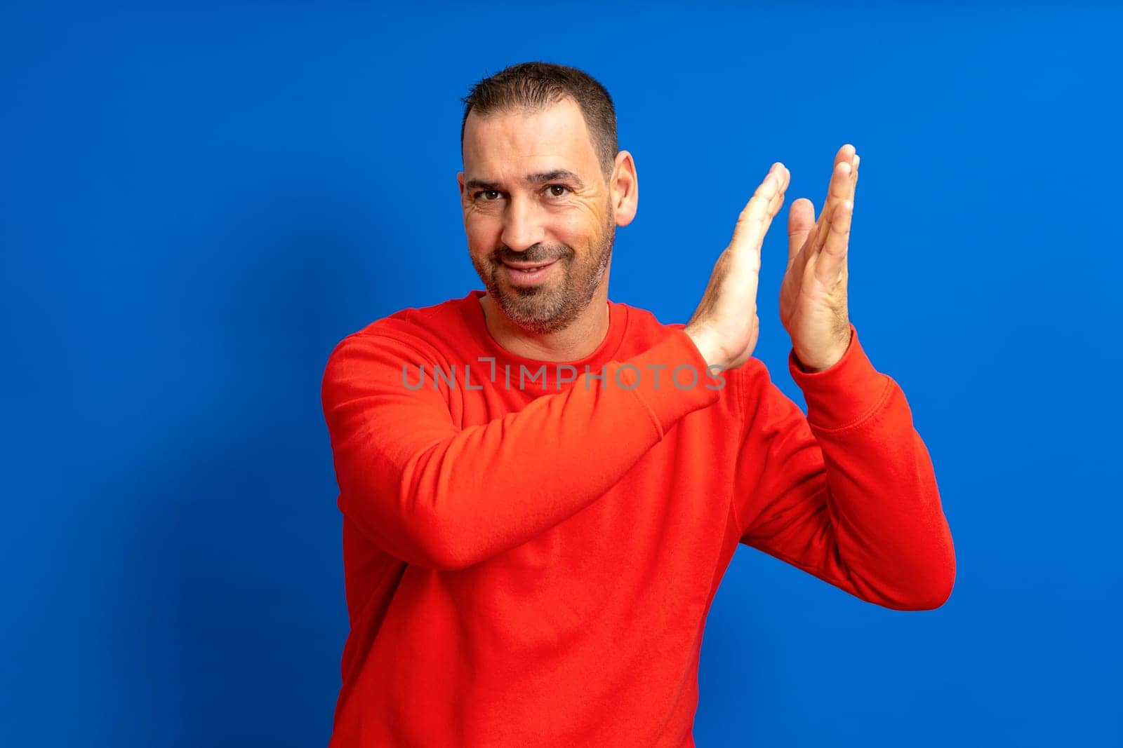Bearded Hispanic man in his 40s wearing a red sweatshirt clapping excited about the great spectacle he just witnessed, isolated over blue background