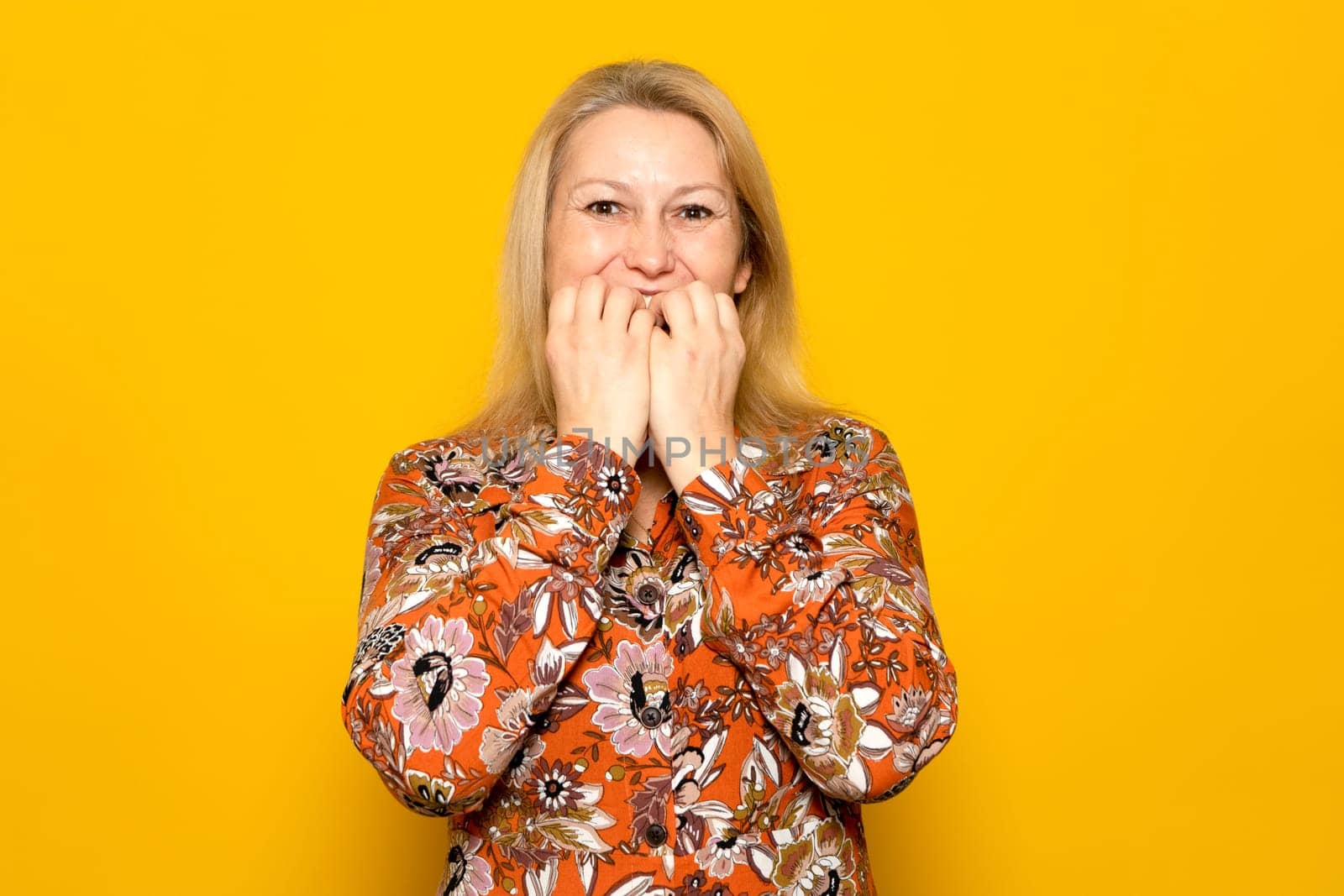 Caucasian blonde woman in her 40s in a patterned dress biting her nails with a nervous gesture, she is in a difficult situation and suffering from anxiety. Isolated on yellow background.
